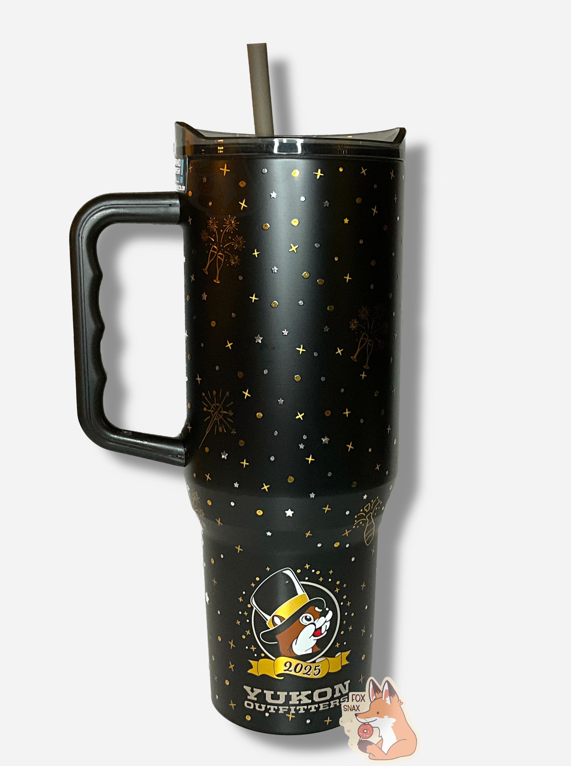 A black, grey, and gold 40 oz tumbler.  The tumbler background is a matte black, and it's streaked with gold and silver dots, gold and silver small cross/x figures, exploding fireworks in gold, and a popping champagne bottle in gold.  The handle is shiny plastic black; the lid is grey/black, partially transparent plastic, and the straw is grey/black as well.  The bottom of the tumbler has a Buc-ee's logo, with Buc-ee the Beaver wearing a black top hat with a gold brim.  It reads 2025, then YUKON OUTFITTERS.