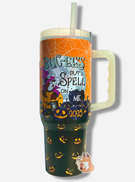 A picture of a tall, orange and black, sorta skinny tumbler, with BUC-EE'S PUT A SPELL ON ME in an old gothic text in black.  There's a lot going on in this tumbler: the background has a full moon, there's Buc-ee dressed as a warlock in purple outfit, in a jack-o-lantern.  Glow in the dark spiderwebs dot the sky.  The tumbler is paint covering stainless steel.  The handle, top, and straw are plastic.  Lit up jack-o-lanterns are piled at the bottom of the tumbler.