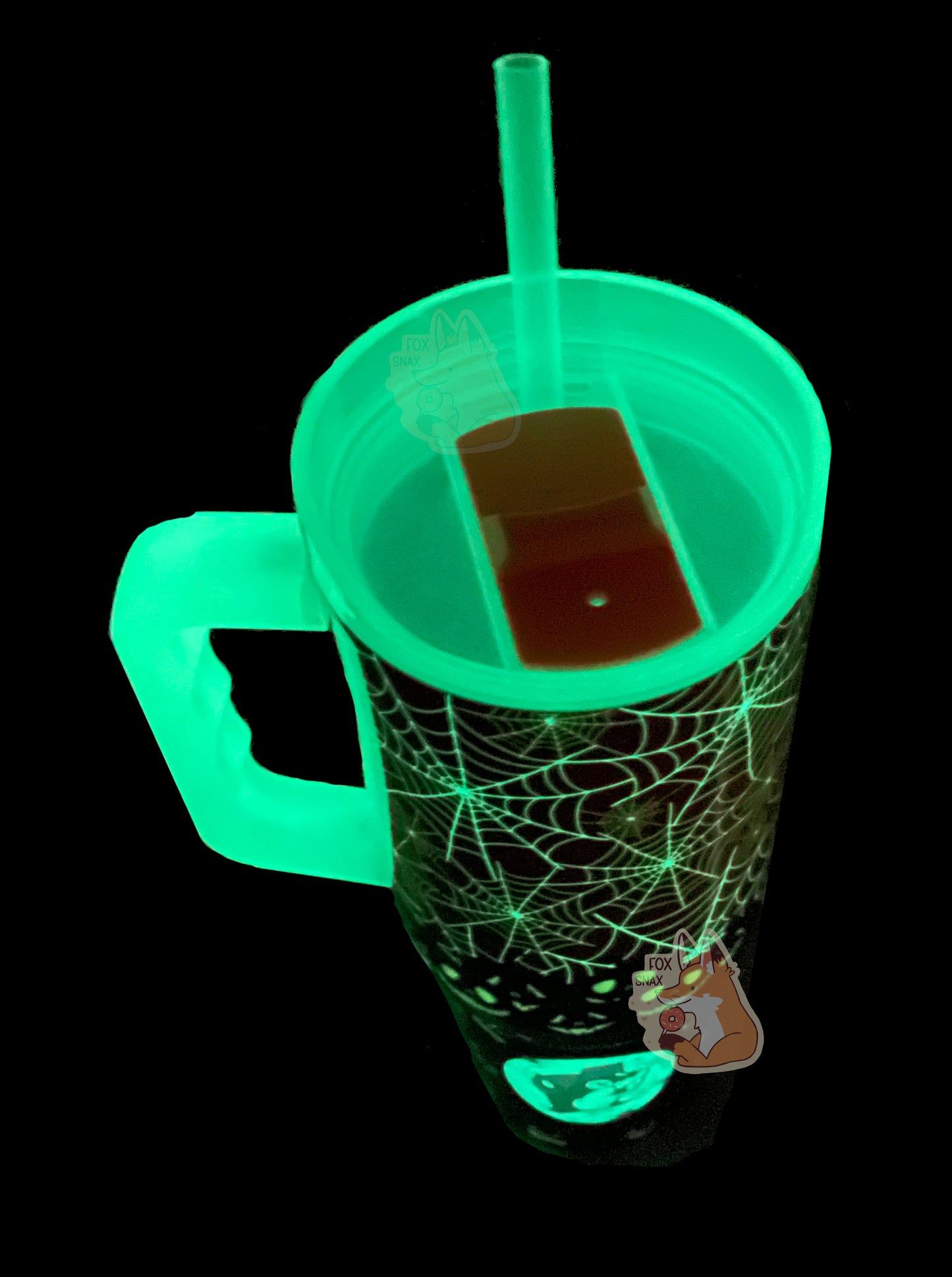 A three-quarters top down view of a tall, dark sorta skinny tumbler.  The spiderwebs on the top of the tumbler are glowing green, as are the jack-o-lanterns on the bottom.  The straw, lid, and handle are also glowing green, illuminating the edges of the slider lid.  On the bottom of the tumbler is a glowing full moon with Buc-ee the Beaver's head in the middle.