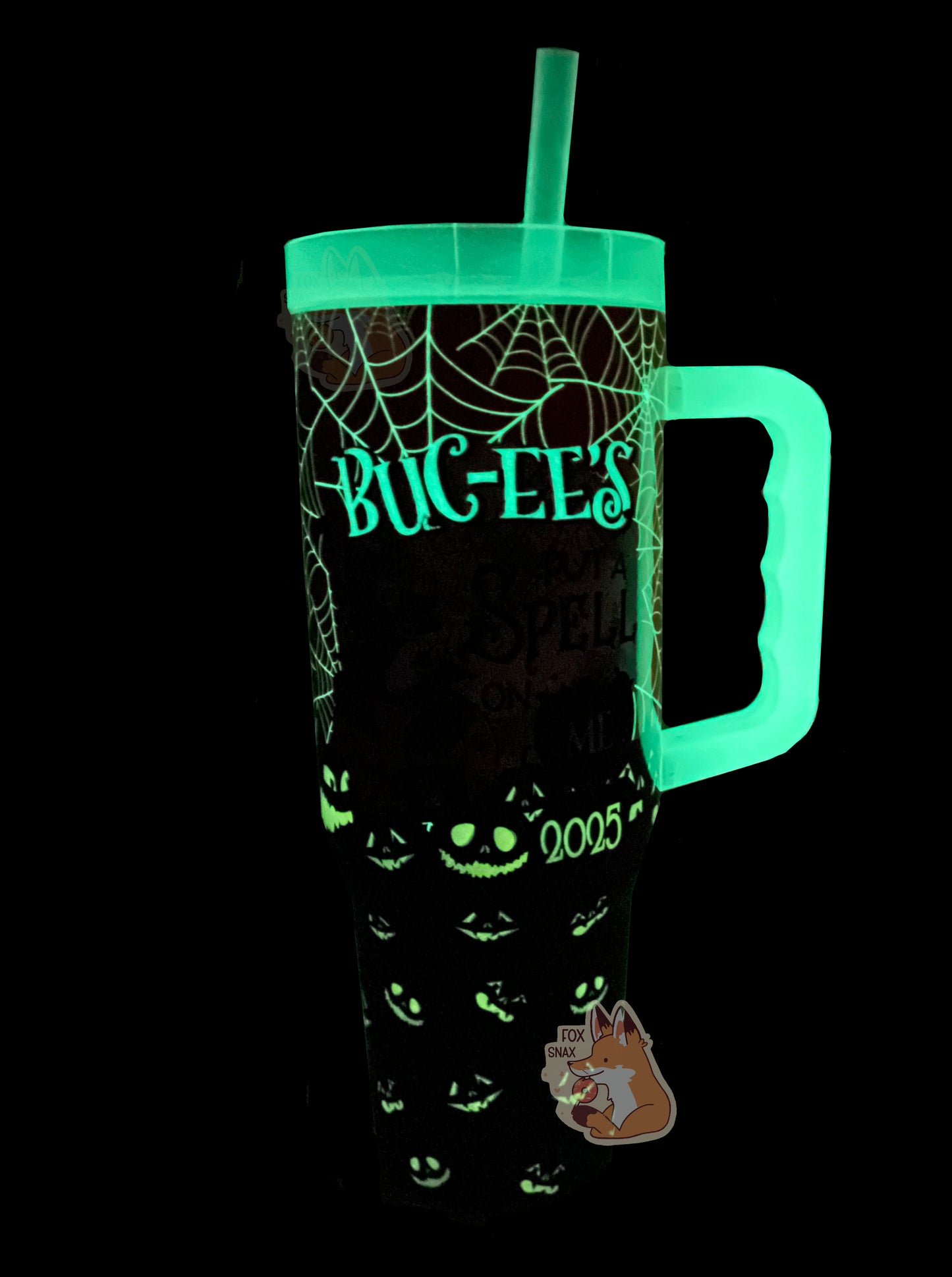 A picture of a tall, sorta skinny tumbler, with BUC-EE'S  in a gothic text. The text, part of the spiderwebs, 2025 logo, and jack-o-lanterns are glowing in the dark on the mug surface.  The straw, lid, and handle are also glowing.