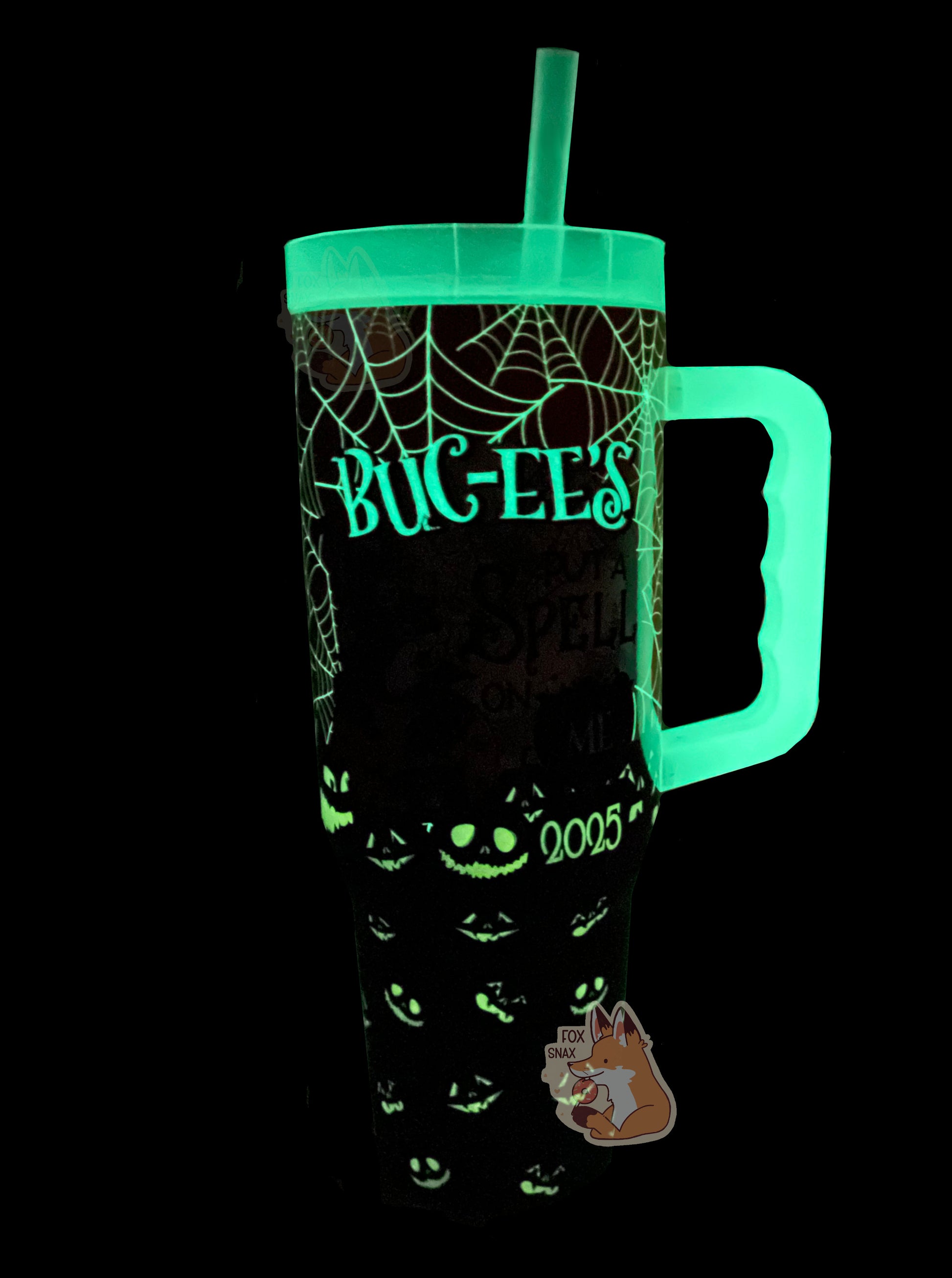 A picture of a tall, sorta skinny tumbler, with BUC-EE'S  in a gothic text. The text, part of the spiderwebs, 2025 logo, and jack-o-lanterns are glowing in the dark on the mug surface.  The straw, lid, and handle are also glowing.