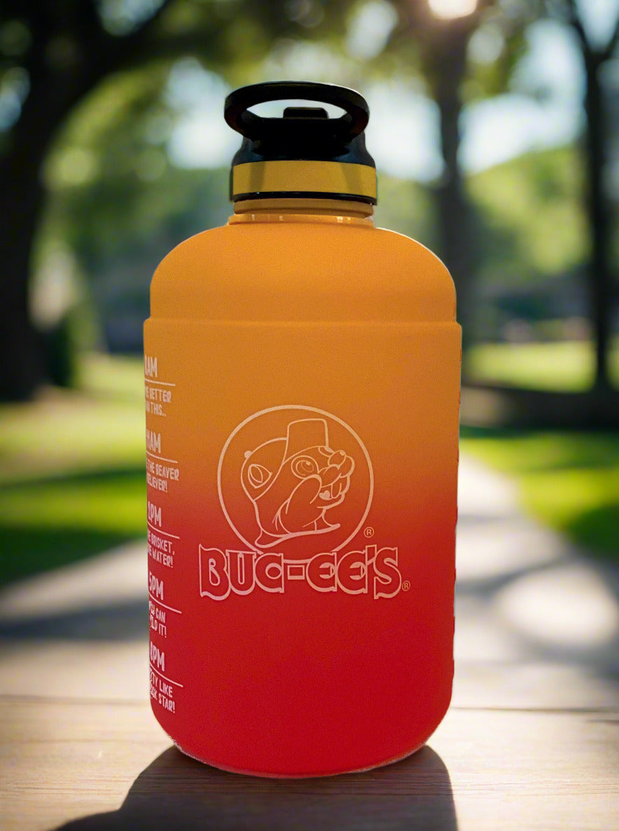A picture of the front of a large, cylindrical hydration jug, made of plastic.  The bottom is a vibrant red, shading upward through a gradient to a brilliant gold at the top.  On the side is a white logo of Buc-ee The Beaver, wearing a ballcap, underneath him: Buc-ee's.  The top of the jug is black plastic, with a yellow band, and a flip-top straw-in-lid in black and yellow.  The jug is sitting on a city park bench.