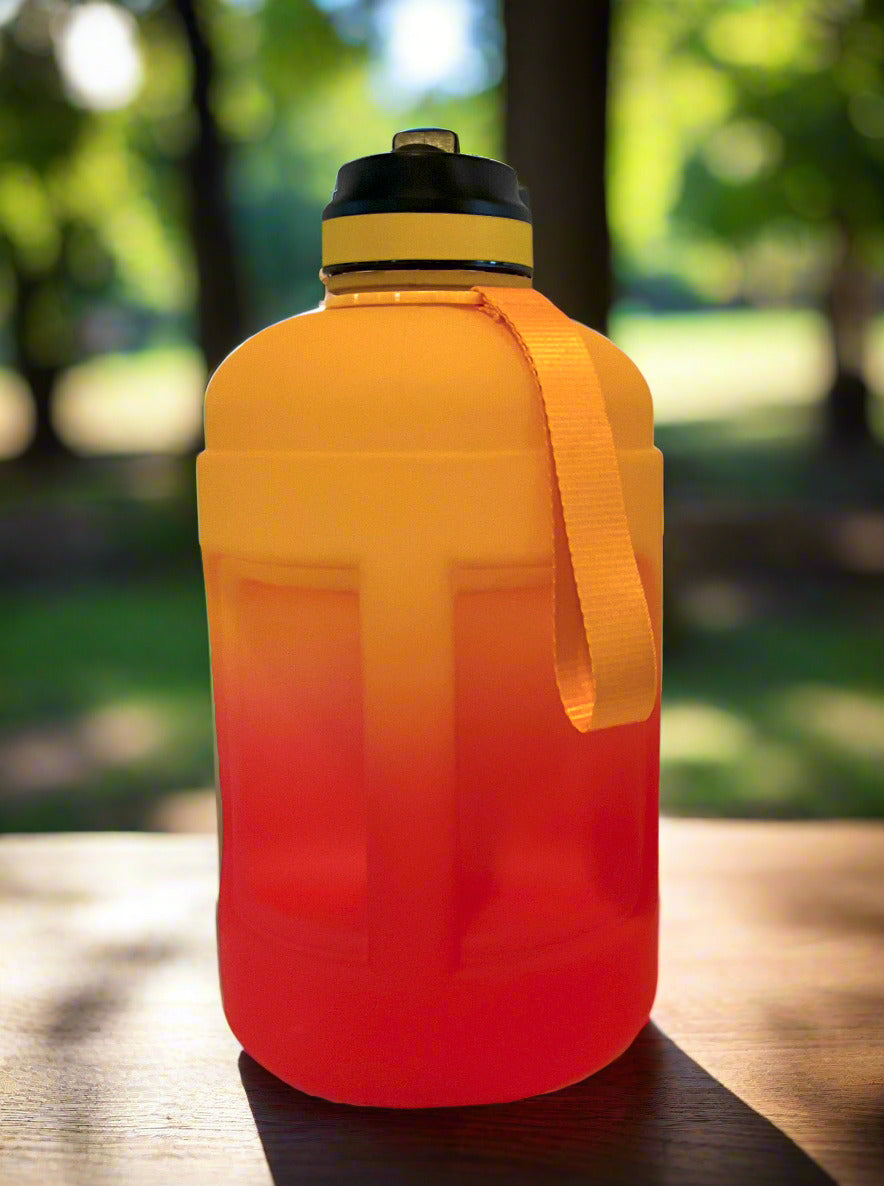 A picture of the back of a large, cylindrical hydration jug, made of plastic.  The bottom is a vibrant red, shading upward through a gradient to a brilliant gold at the top.  A long yellow carry strap is on the side, and on the top, a black plastic handle.  The top of the jug is black plastic, with a yellow band, and a flip-top straw-in-lid in black and yellow.  The back shows a large handle on the side of the jug, in the same gradient colors as the rest of the jug.  The jug is sitting on city park bench.