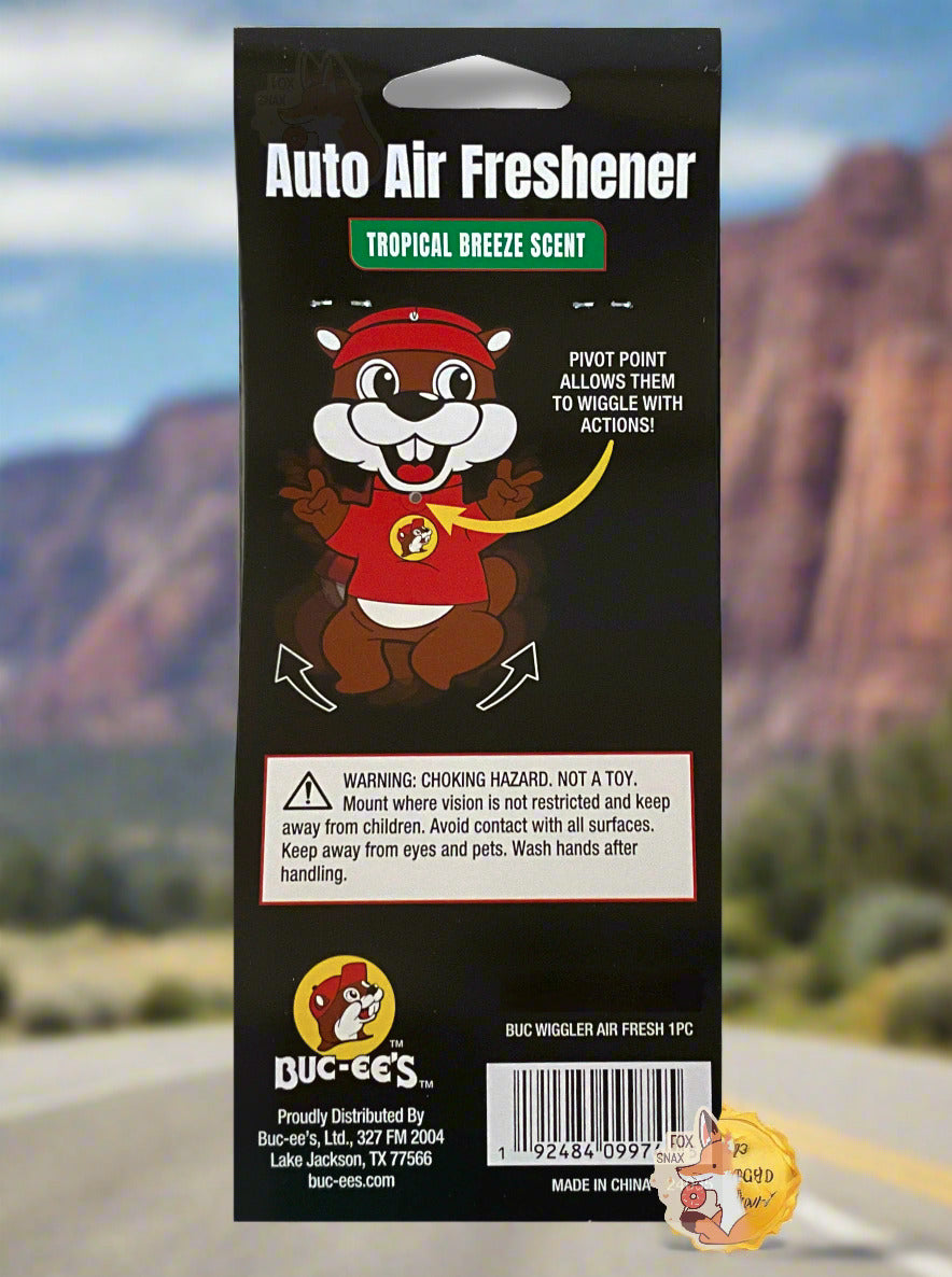 The back of a black package of Buc-ee's Auto Air Freshener, Tropical Breeze Scent, with a picture of Buc-ee The Beaver, with yellow arrows indicating his arms and legs wiggle.  Contains: 1 Air Freshener.  The background is of a beautiful desert highway; several mountains can be seen in the distance, and the grey asphalt of a USA highway is seen on the bottom.