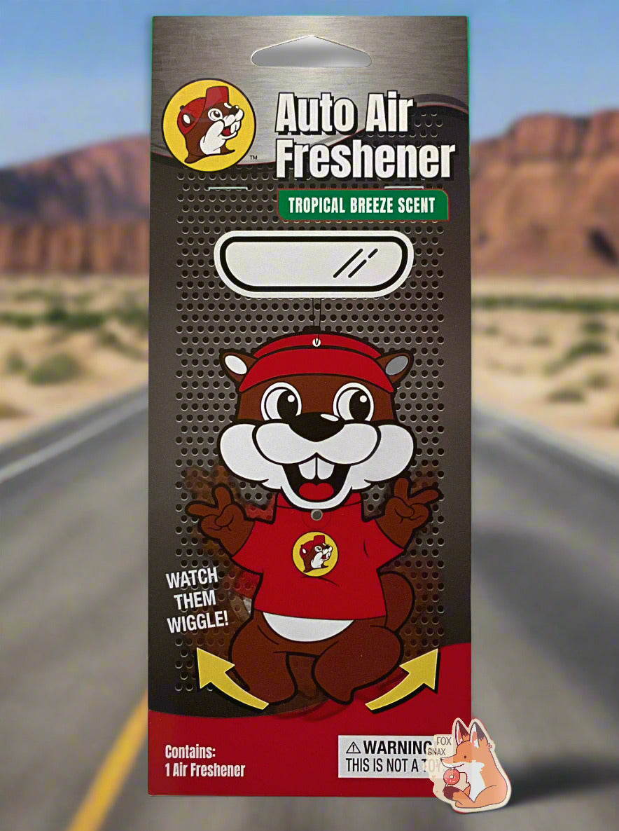The front of a black/grey package of Buc-ee's Auto Air Freshener, Tropical Breeze Scent, with a picture of Buc-ee The Beaver, with yellow arrows indicating his arms and legs wiggle.  Contains: 1 Air Freshener.  The background is of a beautiful desert highway; several mountains can be seen in the distance, and the grey asphalt of a USA highway is seen on the bottom.