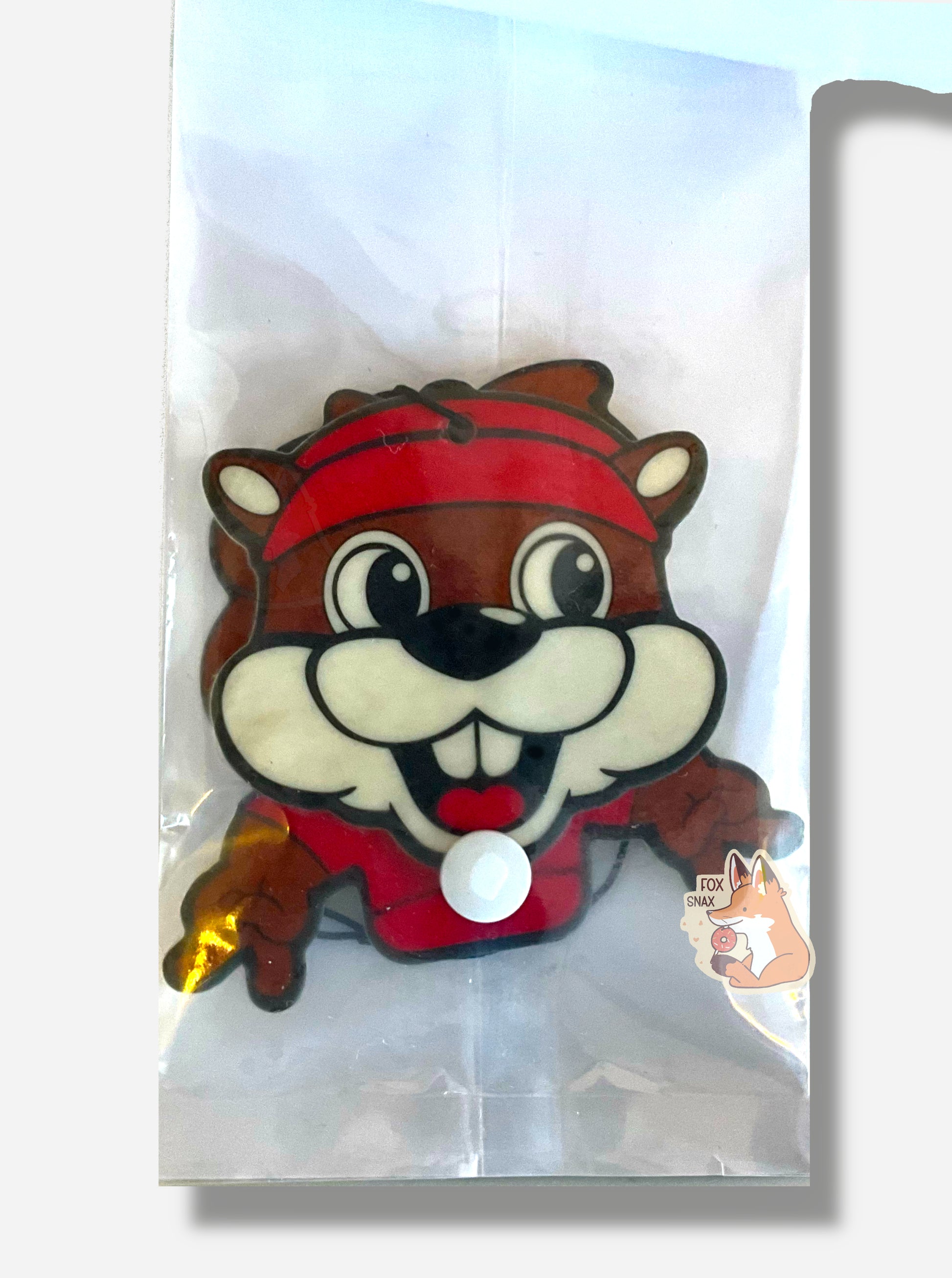 A picture of the inside of a Buc-ee's Wiggler Air Fresher.  Buc-ee is smiling, showing his buck teeth and tongue, and his large eyes are looking to the right.  Each of his hands are holding up the peace sign.