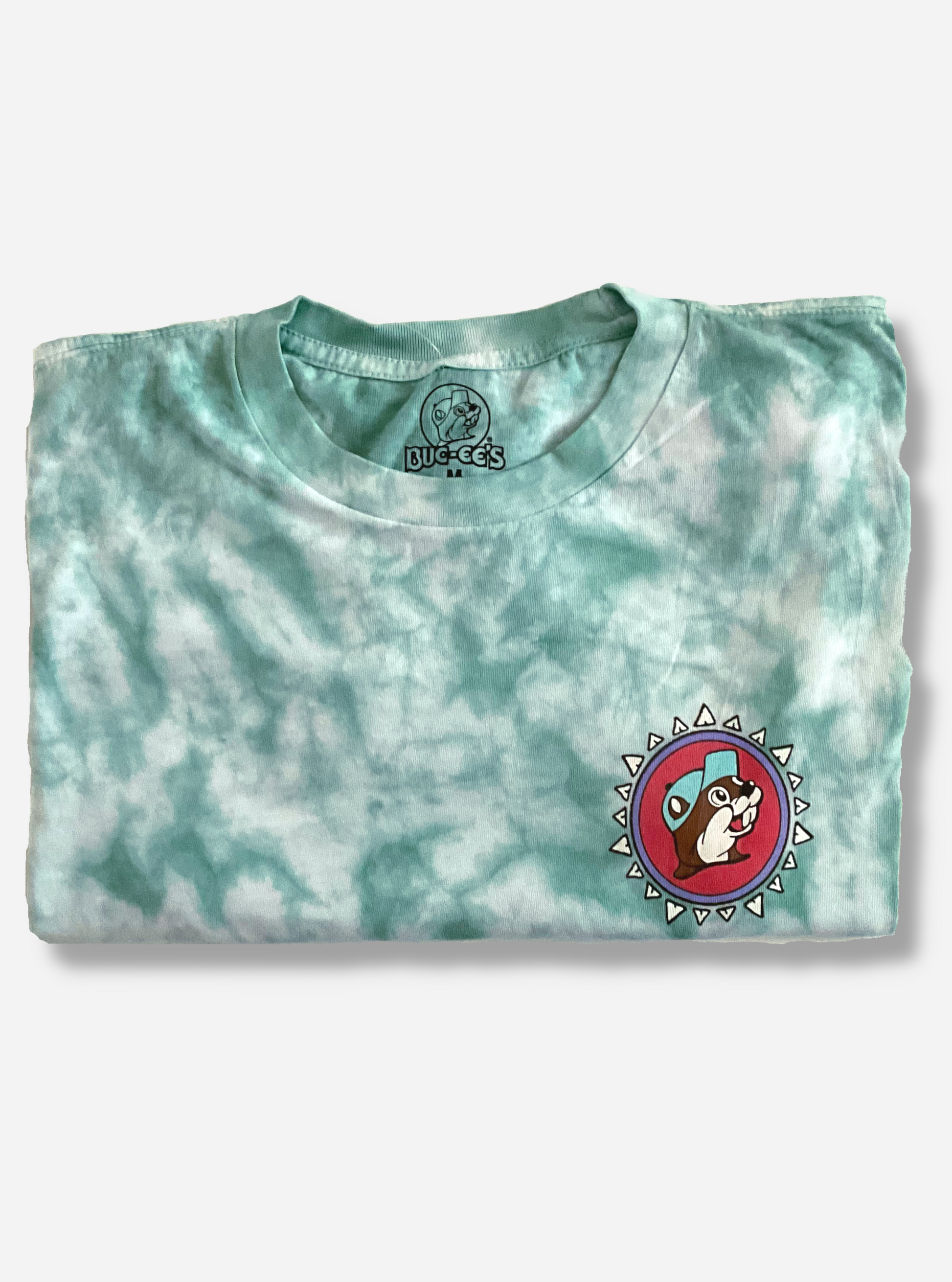 The front of a Buc-ee's Aztec t-shirt.  The background on the shirt is a blue/green tie dye color, against a white background.  On the front right of the shirt is the Buc-ee's logo, but the background is pink, not yellow.  Buc-ee's hat is a light blue, not red.  Finally, the logo is surrounded by a series of white triangles to give it an Aztec-like theme.