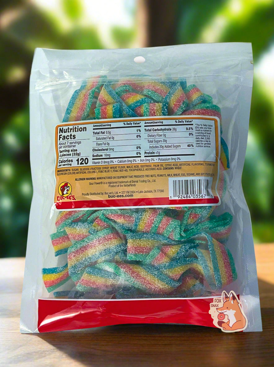 The back of a clear square plastic bag of Buc-ee's Quattro Sour Power Candy Belts.  Inside the bag are a series of rectangular strips of candy, each about an inch and a half long.  The strips are multicolored: green, red, yellow, and blue, and are covered in white sugar.  The background is a solid wooden table with greenery in the background.
