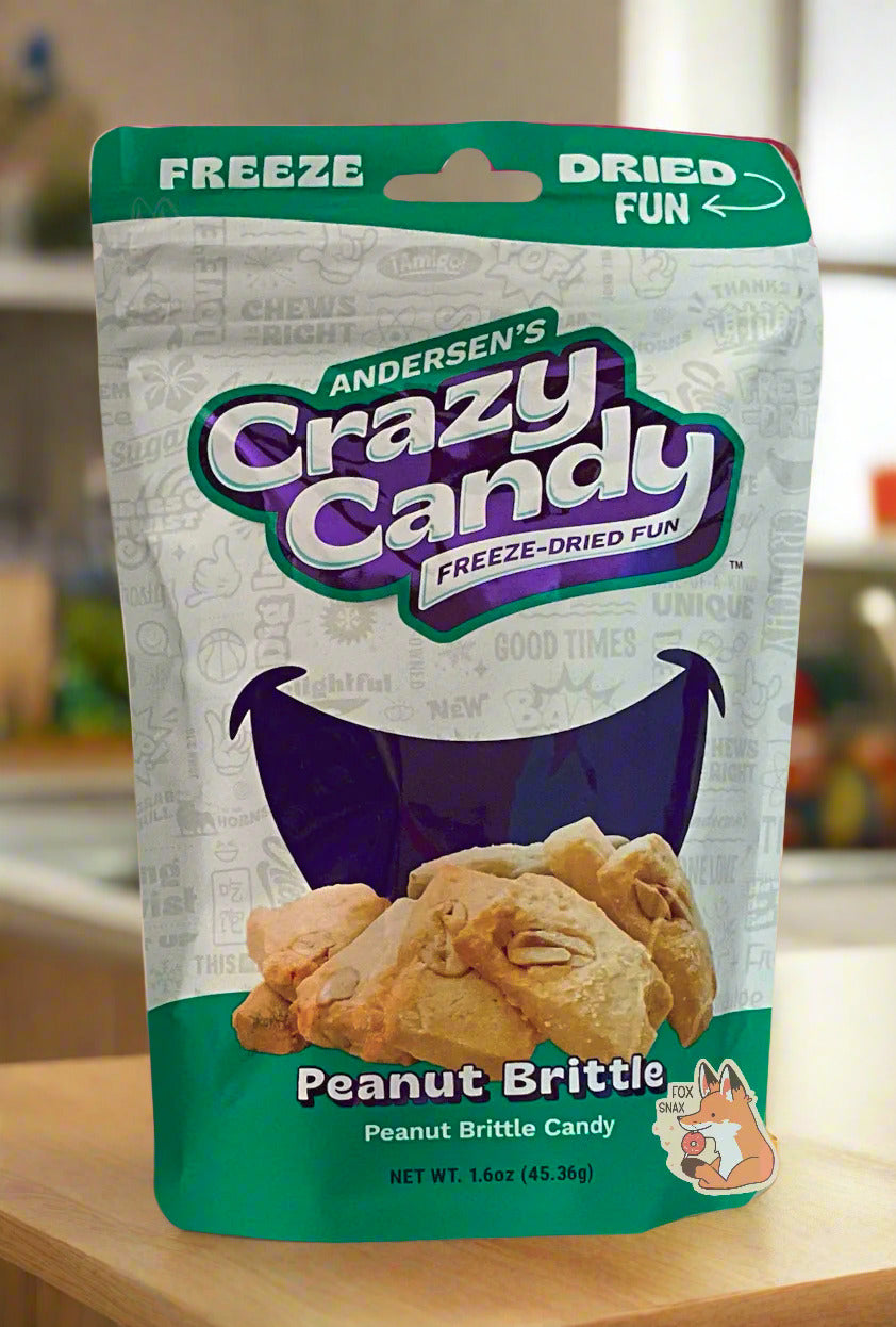 A large bag of Andersen's Peanut Brittle.  The front of the bag also says Peanut Brittle Candy, Net Weight 1.6 oz.  It's Fun, Happy, Candy.  The top and bottom of the bag are green, and the background is a large purple smile against a white background.  Behind the bag is a pretty wood-paneled kitchen; the bag is resting on a light wood countertop.