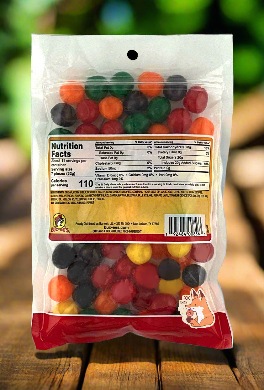 A picture of the back of a resealable bag of Buc-ee's Assorted Sours, which are sour and sweet candies.   The sours inside are spherical, and are multiple shades of red, orange, yellow, green, and purple.  The background is of bright green leafy foliage, with the bag on a wood table.