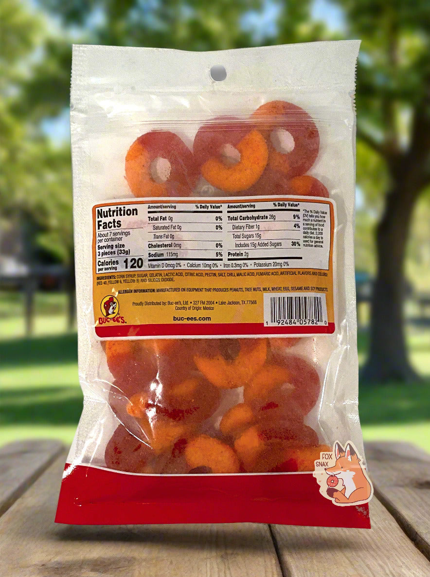 A picture of the back of a clear plastic bag of Buc-ee's Chamoy Peach Rings.  Inside the bag, peach rings can be seen; they are half red and half orange.  The bag is sitting on an outdoor picnic table; peach trees can be seen in the background on a sunny day.