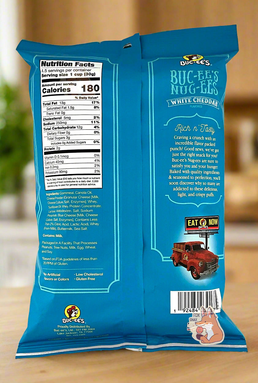 A picture of the back of a opaque, blue and white bag of Buc-ee's White Cheddar Nug-ee's. The bag reads: White Cheddar Flavored, Craving a crunch with incredible flavor packed punch? Good news, we've got just the right snack for you! Buc-ee's Nug-ees(r) are sure to satisfy you and your hunger.  Baked with quality ingredients & seasoned to perfection, you'll soon discover why so many are addicted to these delicious, light, and crispy puffs. The bag is sitting on a wooden countertop in a kitchen.