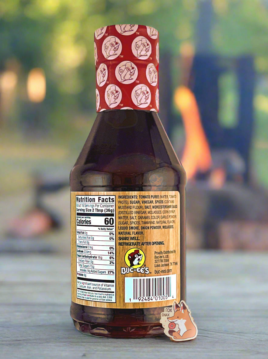 The back of a plastic bottle of Buc-ee's Premium Recipe BBQ Sauce, Original.  The top of the bottle is sealed with a plastic wrap in red and white, dotted with Buc-ee's logos.  The BBQ label is a woodgrain, with a small Buc-ee logo on the bottom.  The label reads: SHAKE WELL.  REFRIGERATE AFTER OPENING.  The bottle is sitting on a wooden table; the outline of a bbq grill with large flames can be seen in the background.