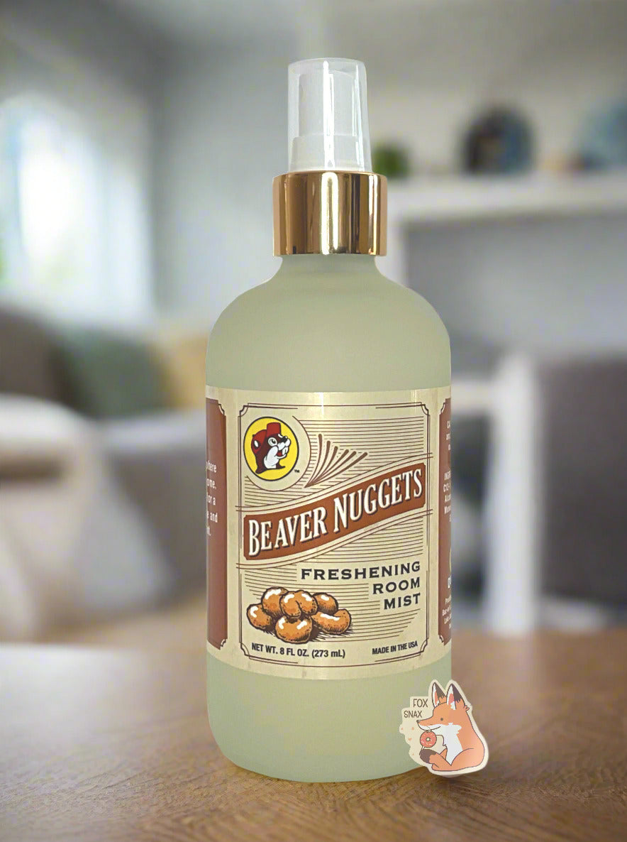 Front of a spray bottle of Buc-ee's Beaver Nuggets Freshening Room Mist on a white background.  The bottle is frosted glass with a white sprayer and gold label, and has the Buc-ee's logo in the top left.  The rest of the label has a series of brown lines, and reads BEAVER NUGGETS, then FRESHENING ROOM MIST, then NET WT 8 FL OZ, then MADE IN THE USA.  The background shows a clean living room, with a grey couch, end tables, and a fireplace.