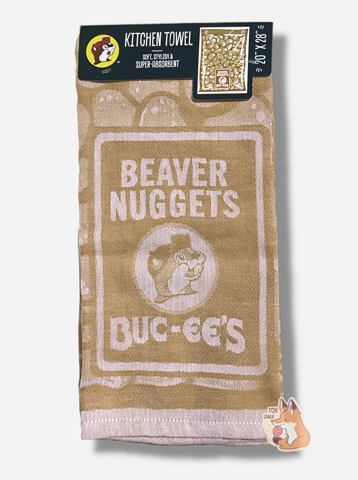 A picture of an 20x28, tan kitchen towel, adorned with BEAVER NUGGETS text, next to a logo of Buc-ee the Beaver.  The full unwrapped towel shows a large rectangular package of Beaver Nuggets.  The top of the towel has a cardboard tag that reads KITCHEN TOWEL, then SOFT, STYLISH & SUPER-ABSORBENT, then 20x28.