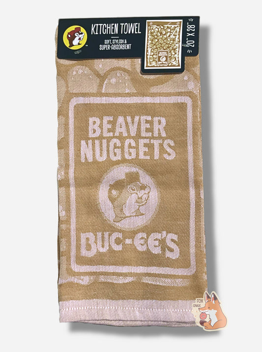 A picture of an 20x28, tan kitchen towel, adorned with BEAVER NUGGETS text, next to a logo of Buc-ee the Beaver.  The full unwrapped towel shows a large rectangular package of Beaver Nuggets.  The top of the towel has a cardboard tag that reads KITCHEN TOWEL, then SOFT, STYLISH & SUPER-ABSORBENT, then 20x28.