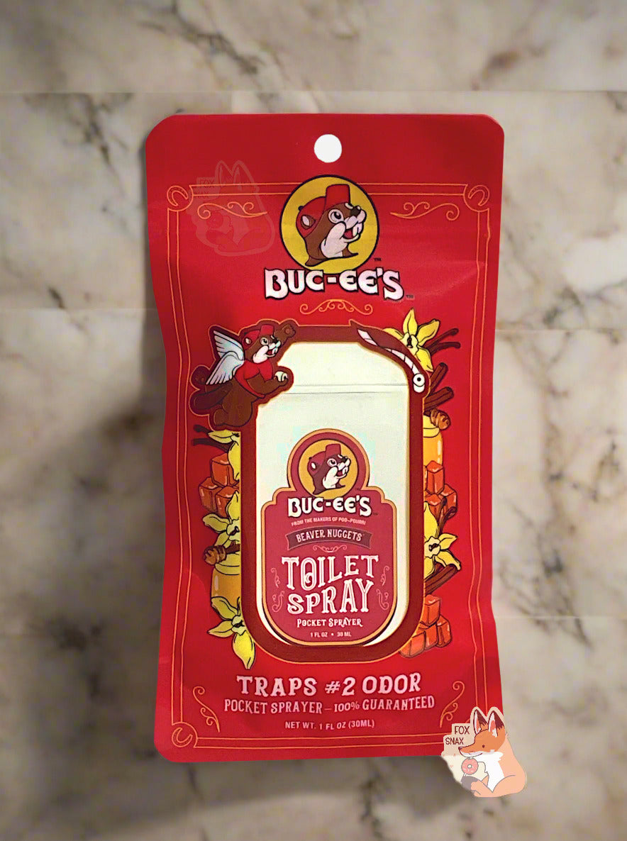Red package of Buc-ee's toilet spray on a white background.  The label, amusingly, in the top left, has Buc-ee with a pair of angel wings, holding a roll of toilet paper in one paw, and in the other, outraised, throwing a roll of toilet paper.  The label reads: BUC-EE'S, FROM THE MAKERS OF POO~POURRI, BEAVER NUGGETS POCKET TOILET SPRAYER, then TRAPS #2 ODOR, then 1 FL OZ.  The background is of tile in a marble bathroom.