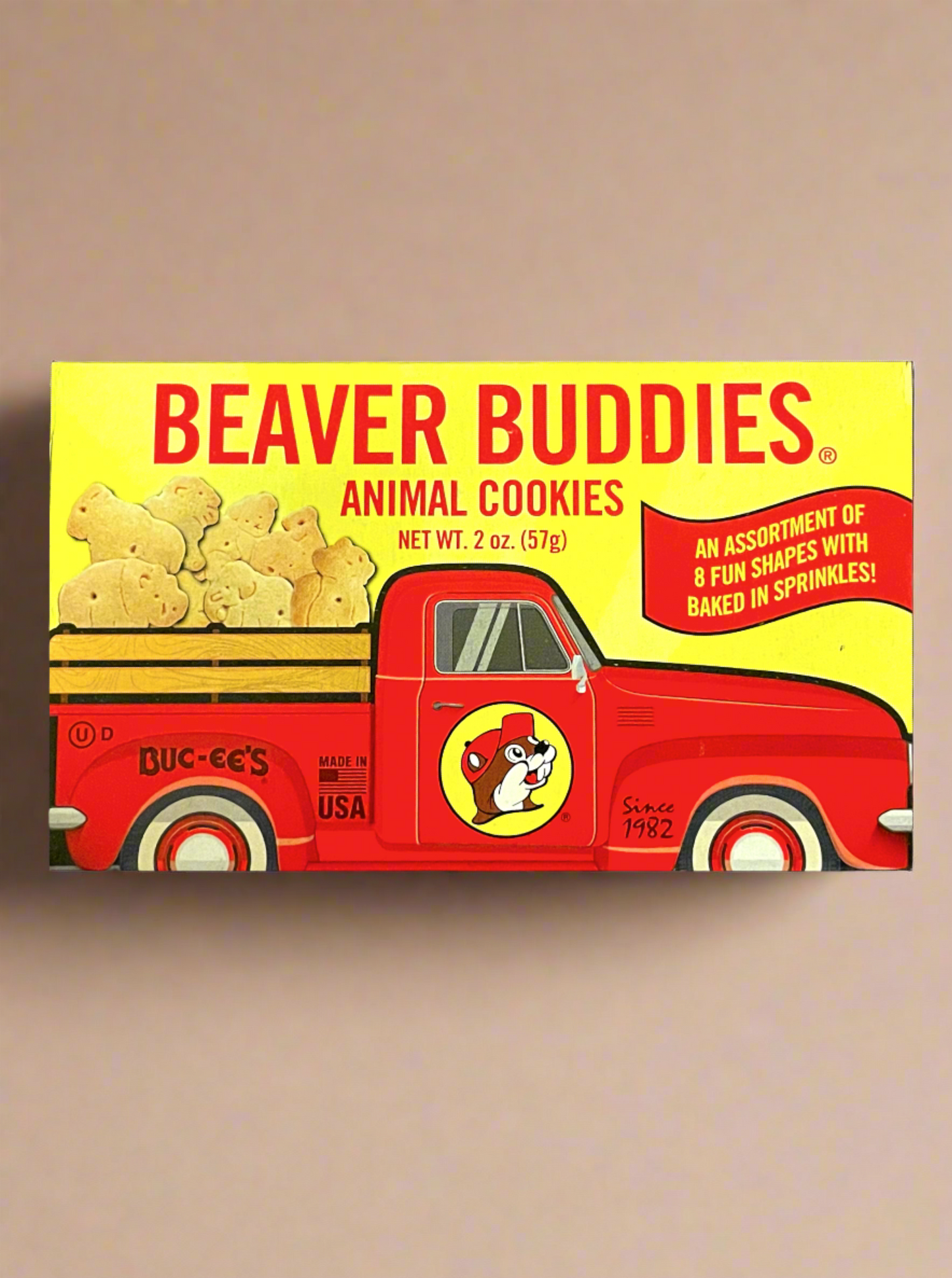 The front of a small rectangular cardboard package of Buc-ee's Beaver Buddies Animal Cookies, sitting on a light tan background. The label has a picture of a red truck with the Buc-ee The Beaver logo on the door panel, and a series of animal cookies in the bed of the truck.  The text reads BEAVER BUDDIES ANIMAL COOKIES, NET WT 2 oz (57g).  AN ASSORTMENT OF 8 FUN SHAPES WITH BAKED IN SPRINKLES!  Made in the USA.