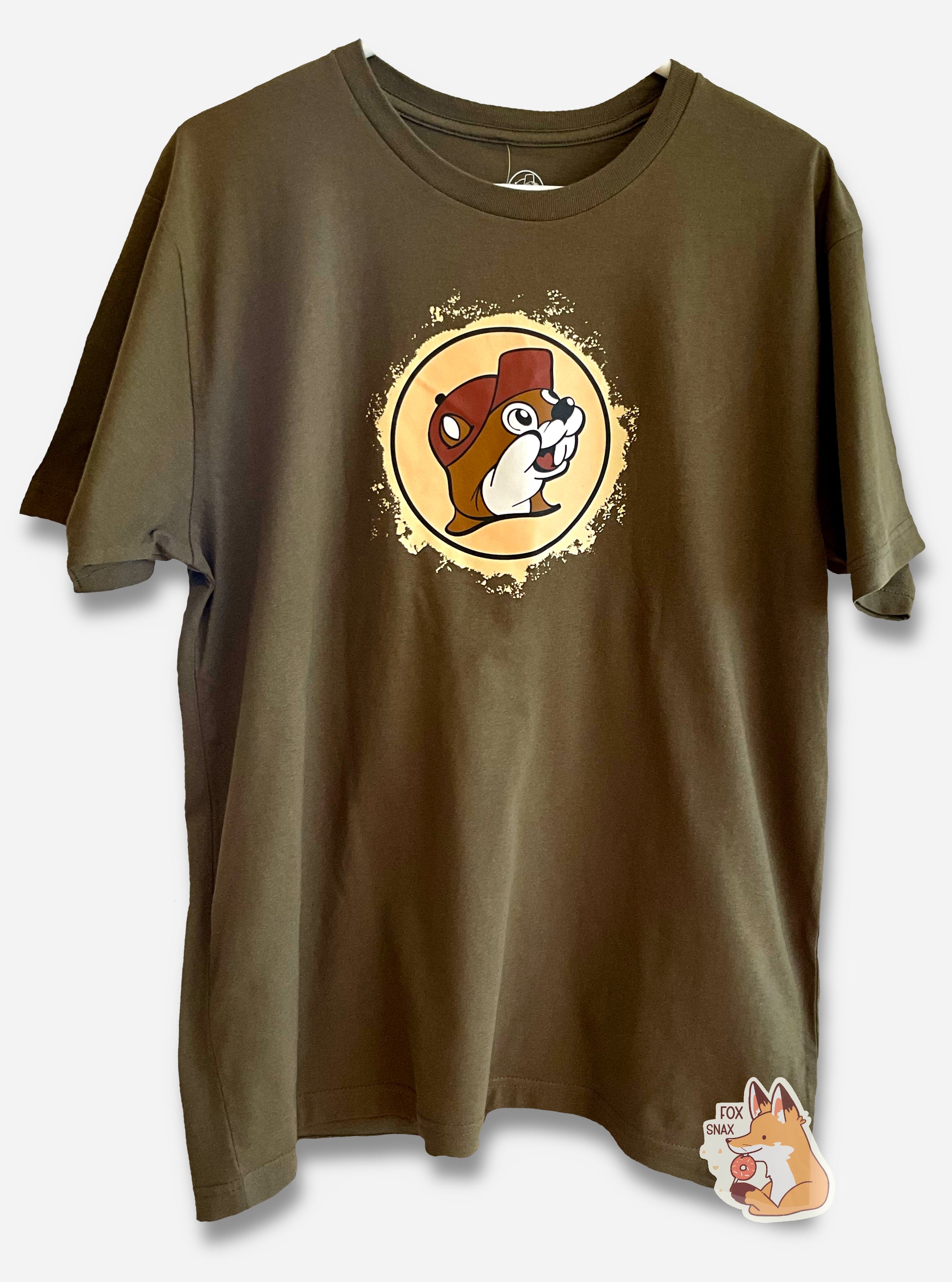 A picture of a tan t-shirt, with the Buc-ee the Beaver logo in the middle.  Surrounding the logo is a beat-up, faded yellow/tan circle, as if the logo itself has been dug up and, previously, subjected to a lot of historical events, as it shows wear.