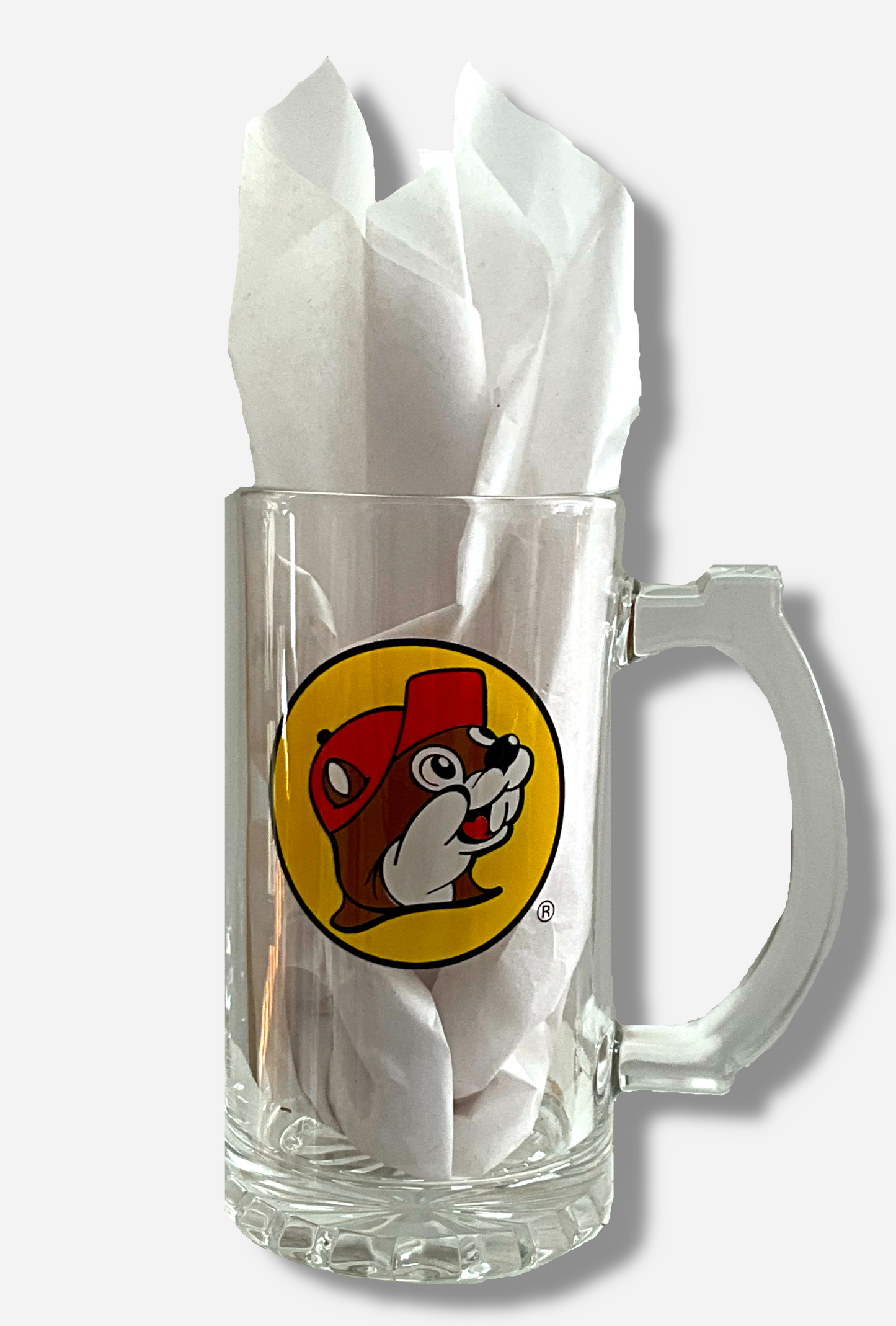 A picture of the front of clear beer mug. The glass is clear. In the middle of the front is the Buc-ee's logo, which appears inside a yellow circle; it is a cheerful beaver wearing a red ballcap, against a yellow circle background.  The handle of the beer mug is also clear, and slightly blocky.  A ridged thumb area on the top can be seen.  The mug is filled with a white tissue paper to better illustrate what it looks like with material in it.