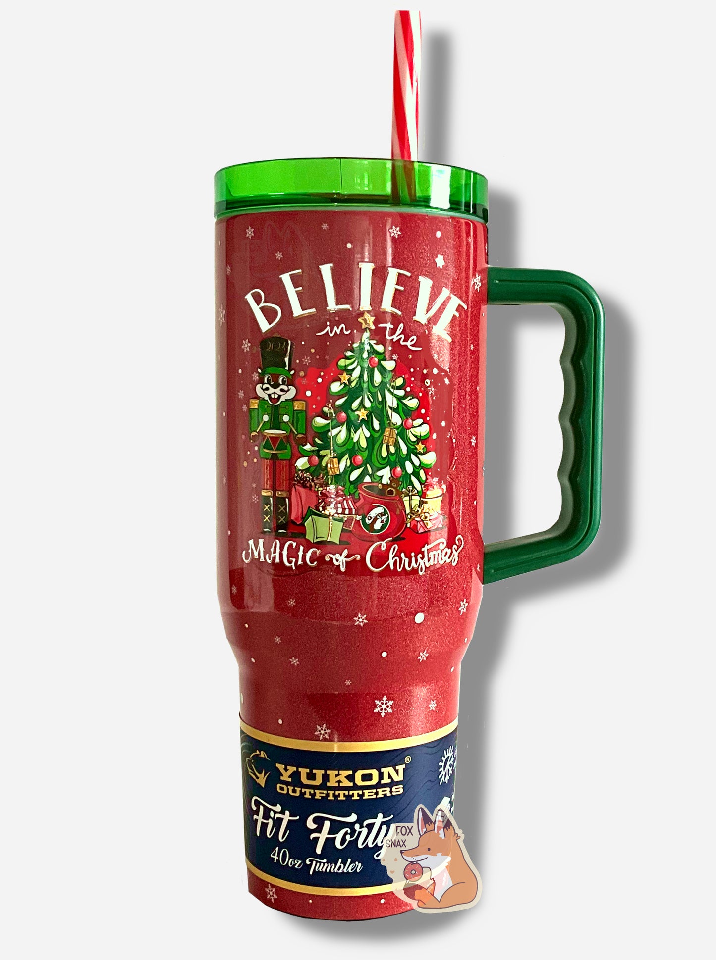 A picture of a tall, maroon and red, skinny tumbler, with Believe In the Magic of Christmas written in white text. In this tumbler, the background has a Christmas tree with presents aplenty below, Buc-ee is standing to the left, wearing a green nutcracker outfit, and the background is covered in white snowflakes. The tumbler is paint covering stainless steel.  The handle, top, and candy-cane colored straw are plastic. A paper wrapper around the bottom reads: Yukon Outfitters.  Fit Forty 40 oz Tumbler.  