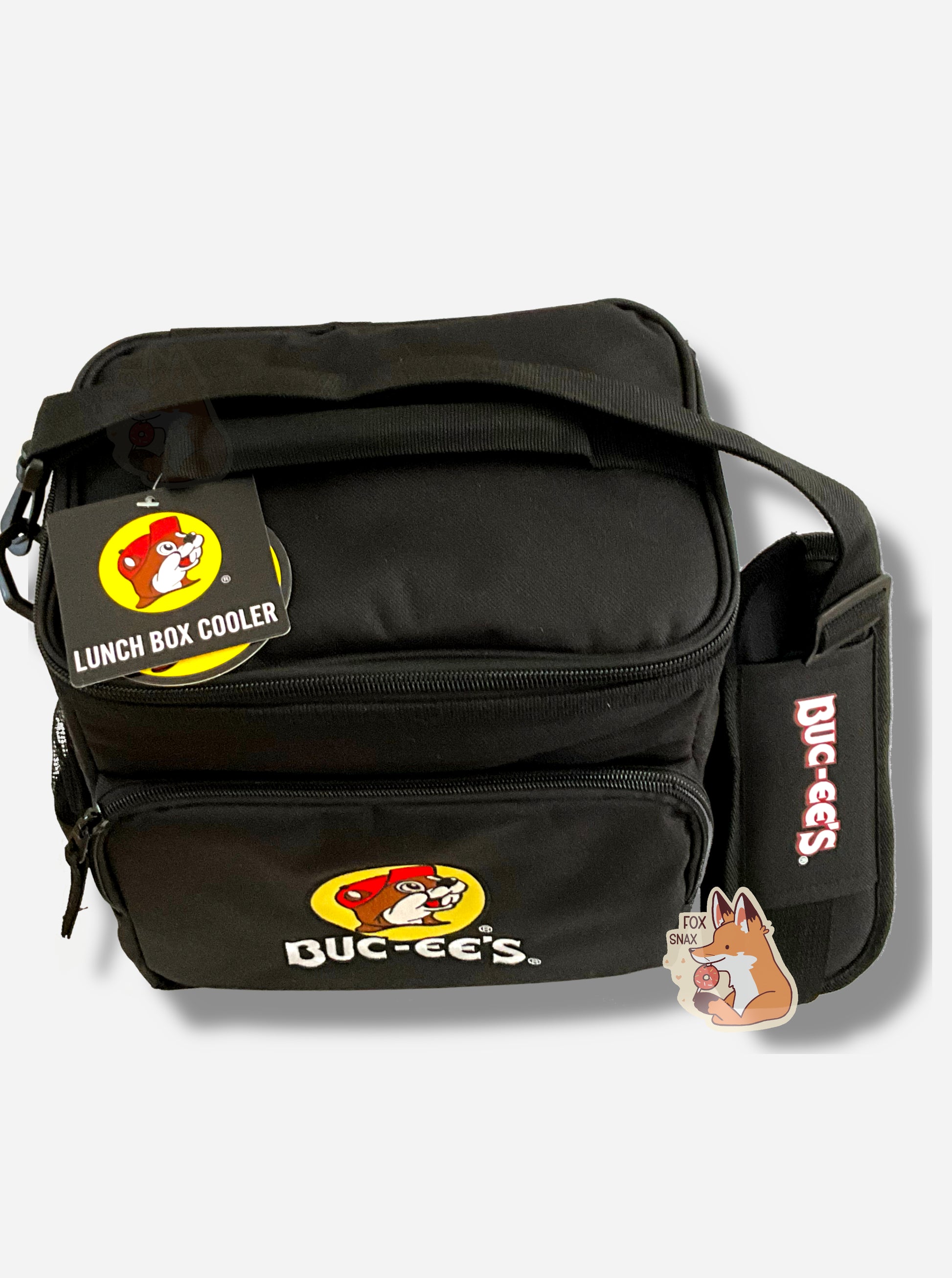 A picture of a three-quarters view of the front of a large lunch box cooler.  The outer surface is made of black polyester, and has a large black strap on it that reads BUC-EE'S.  The front of the lunch box has a zip pouch, with the Buc-ee's logo on it: a grinning beaver wearing a red hat against the background of a yellow circle.  The sides of the tote has mesh pouches, the top sealed by a zipper.