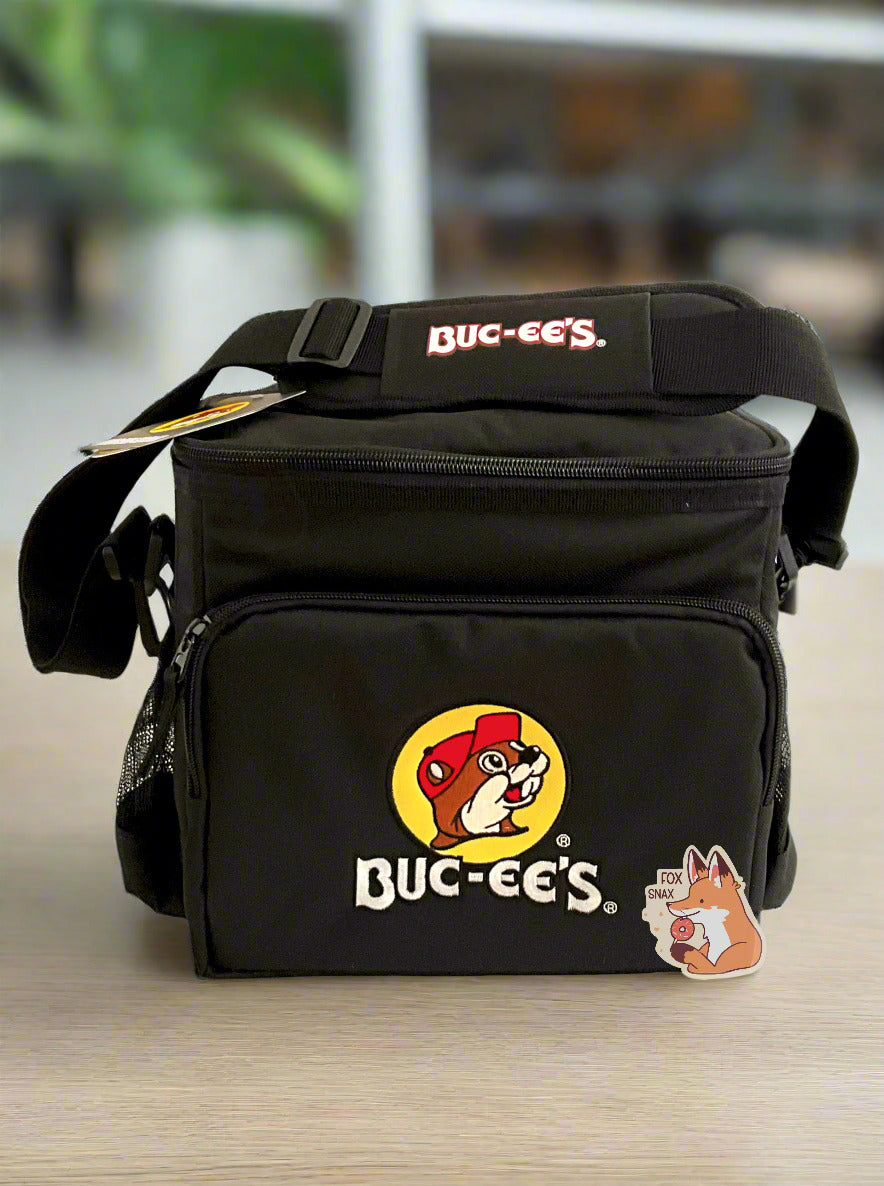 A picture of a three-quarters view of the front of a lunch box cooler.  The outer surface is made of black polyester, and has a large black strap on it that reads BUC-EE'S.  The front of the lunch box has a zip pouch, with the Buc-ee's logo on it: a grinning beaver wearing a red hat against the background of a yellow circle.  The sides of the tote has mesh pouches, the top sealed by a zipper.  The lunchbox is sitting on a pretty light wood table with plants and a work cafeteria in the background.