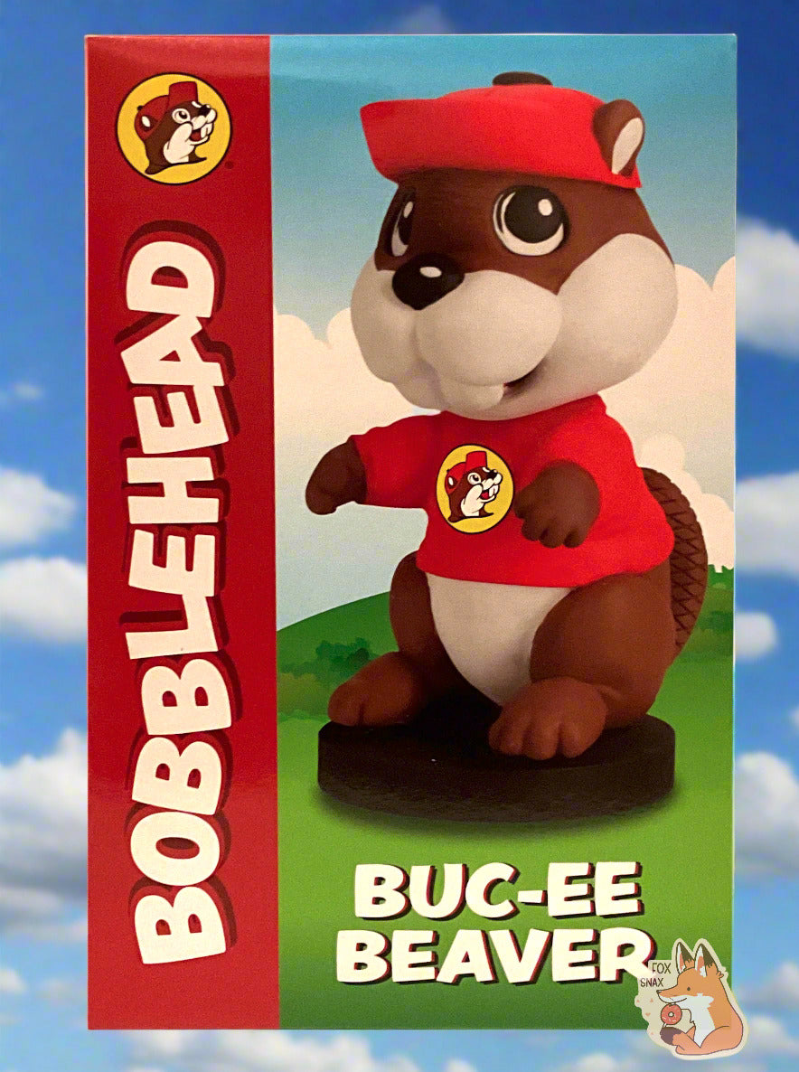 A picture of the front of a box for a Buc-ee The Beaver Bobblehead.  It shows an adorable plastic beaver with a big fat beaver head; Buc-ee is wearing his usual red ball cap and Buc-ee's shirt. and has his arms in front of his body.  He is standing on a small circular black pedestal; his tail can be seen behind him.  The background is of a green cartoon meadow behind the bobblehead; the background behind the box is of a blue sky with fluffy white clouds.