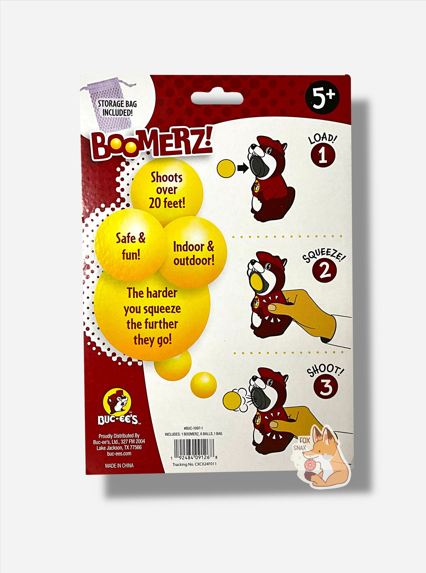 The back of a package of Buc-ee's Boomerz!  The top left reads STORAGE BAG INCLUDED, and shows a small white mesh bag.  The top right reads 5+.  A three-step diagram showing LOAD!, where a yellow ball is placed in Buc-ee's mouth, then SQUEEZE!, with a hand squeezing the Buc-ee's toy's side, then SHOOT!, where the ball pops out of Buc-ee's mouth with a gust of wind.  The rest of the text reads SHOOTS OVER 20 FEET! SAFE & FUN! INDOOR & OUTDOOR, and THE HARDER YOU SQUEEZE THE FURTHER THEY GO!