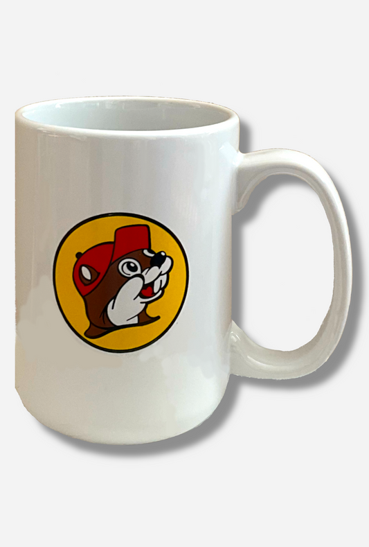 A picture of a white ceramic coffee mug, with a white handle.  The middle of the mug has the Buc-ee's logo on it, which is a picture of a grinning beaver head with brown and white fur, wearing a red ballcap.  The background behind the beaver is a yellow circle, outlined in black.