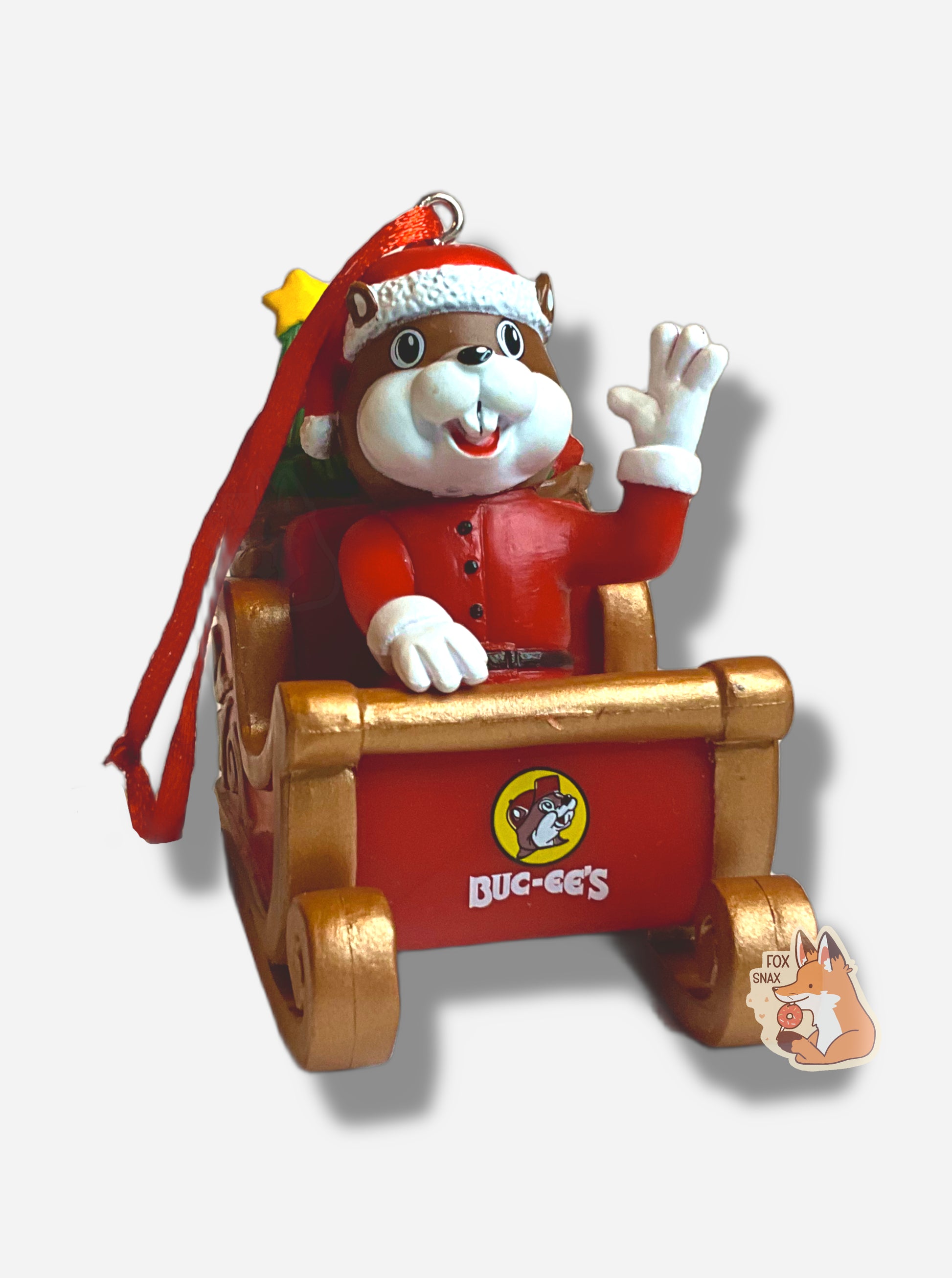 A picture of the front of a bright, plastic, Christmas ornament.  The ornament is of Buc-ee the Beaver -  a smiling beaver wearing a red Christmas stocking cap and Santa outfit.  Buc-ee is riding in a gold sleigh, with the Buc-ee's logo prominently in the front.  His left arm is raised in a greeting.  A red ribbon tied to a very small  hook is poking out of Buc-ee's head.