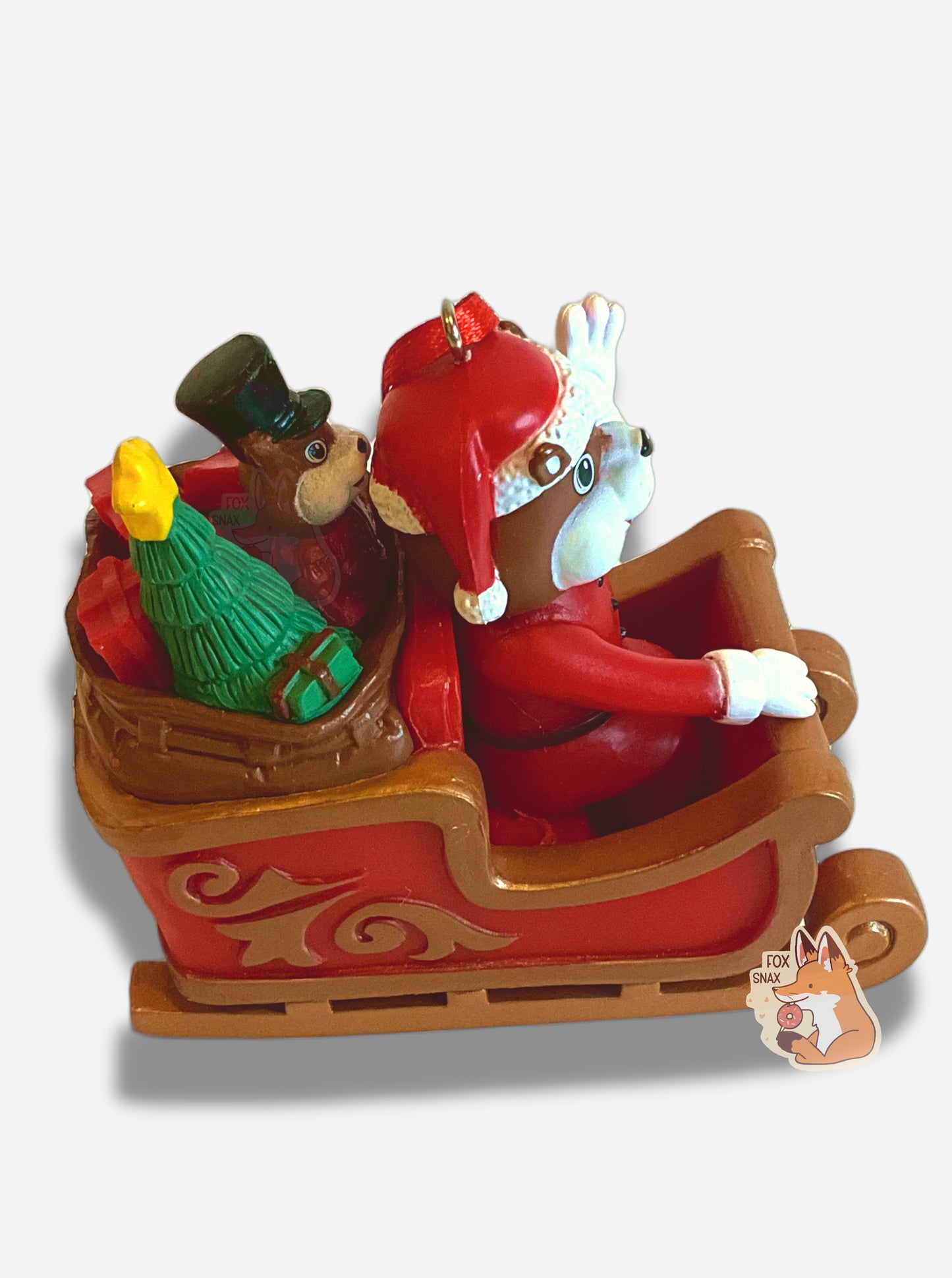 A picture of the side of a bright, plastic, Christmas ornament.  The ornament is of Buc-ee the Beaver -  a smiling beaver wearing a red Christmas stocking cap and Santa outfit.  Buc-ee is riding in a gold and red sleigh; his left arm is raised in a greeting.  A red ribbon tied to a very small  hook is poking out of Buc-ee's head.  In the back of the sleigh are a green Christmas tree with a yellow star, a beaver "nutcracker", and  brightly colored present boxes.