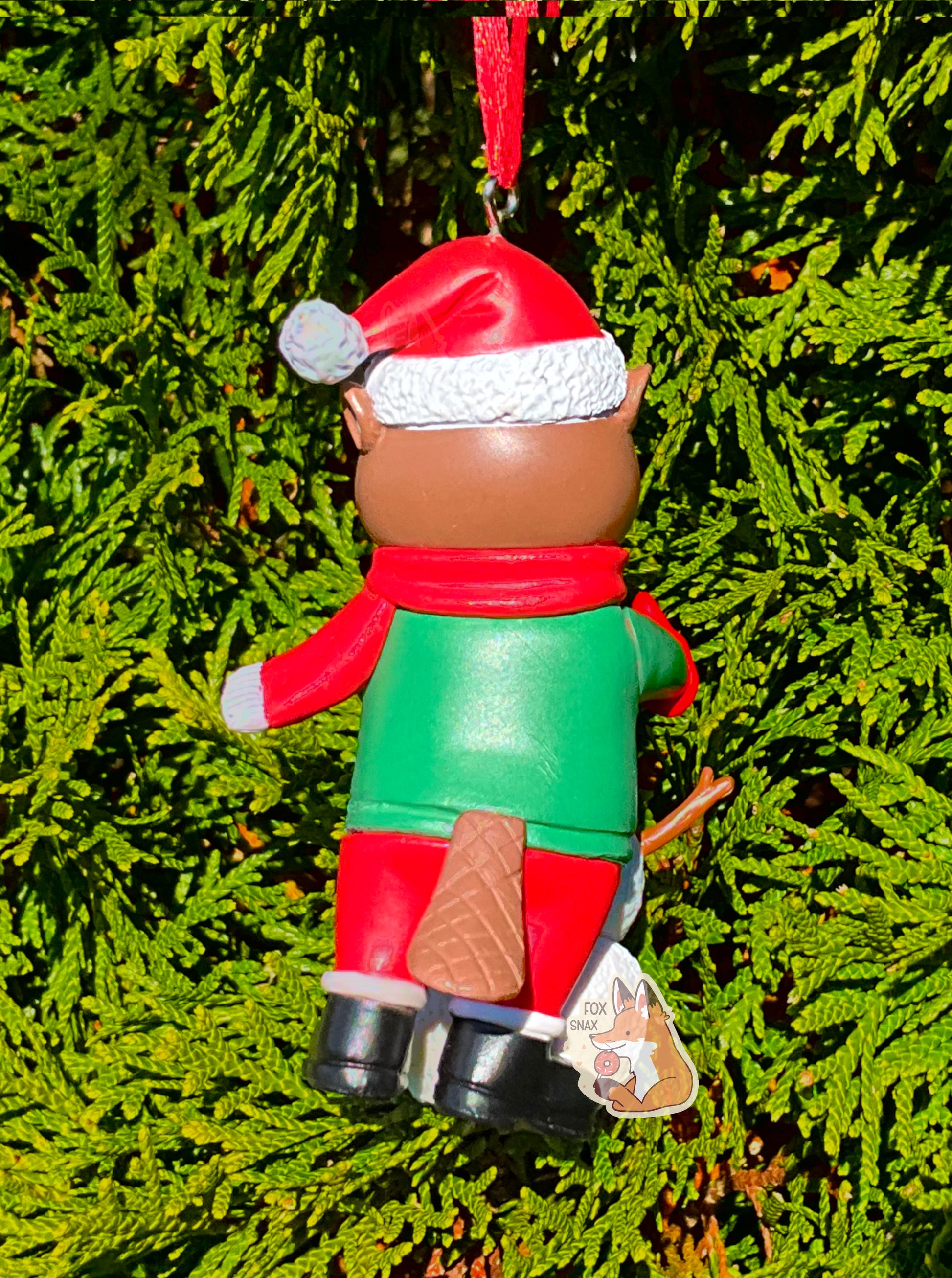 A picture of the back  of a bright, plastic, Christmas ornament.  The ornament is of Buc-ee's the Beaver -  a smiling beaver wearing a red Christmas stocking cap. Buc-ee is wearing a red scarf and green coat, with red mittens and Santa pants and black boots.  His brown-furred beaver tail is poking out of the back of his pants.  A red ribbon pokes out from the top of the ornament's body, tying it to the green evergreen branches of a pretty tree in the background.