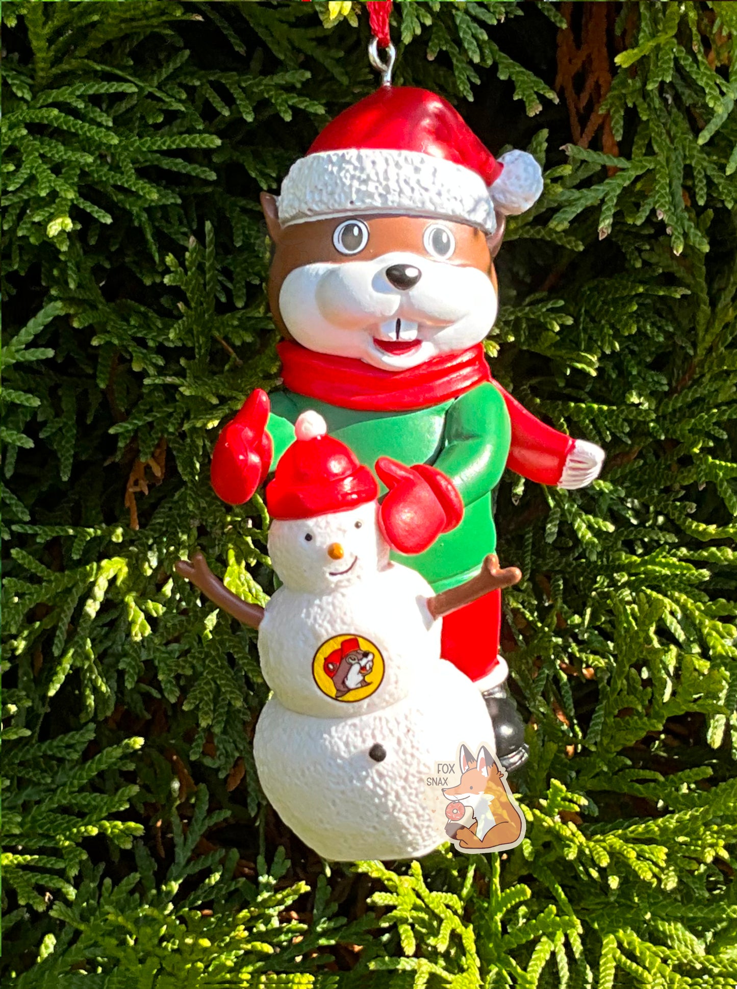 A picture of the front of a bright, plastic, Christmas ornament.  The ornament is of Buc-ee's the Beaver -  a smiling beaver wearing a red Christmas stocking cap. Buc-ee is wearing a red scarf and green coat, with red mittens and Santa pants and black boots.  In front of Buc-ee is a small snowman, with the Buc-ee's logo on it's chest.  A red ribbon pokes out from the top of the ornament's body, tying it to the green evergreen branches of a pretty tree in the background.