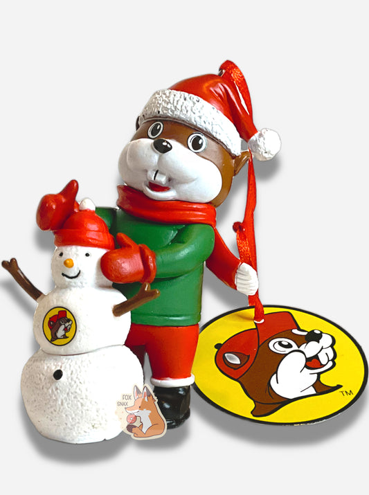 A picture of the front of a bright, plastic, Christmas ornament.  The ornament is of Buc-ee's the Beaver -  a smiling beaver wearing a red Christmas stocking cap. Buc-ee is wearing a red scarf and green coat, with red mittens and Santa pants and black boots.  In front of Buc-ee is a small snowman, with the Buc-ee's logo on it's chest.  A red ribbon pokes out from the top of the ornament's body.