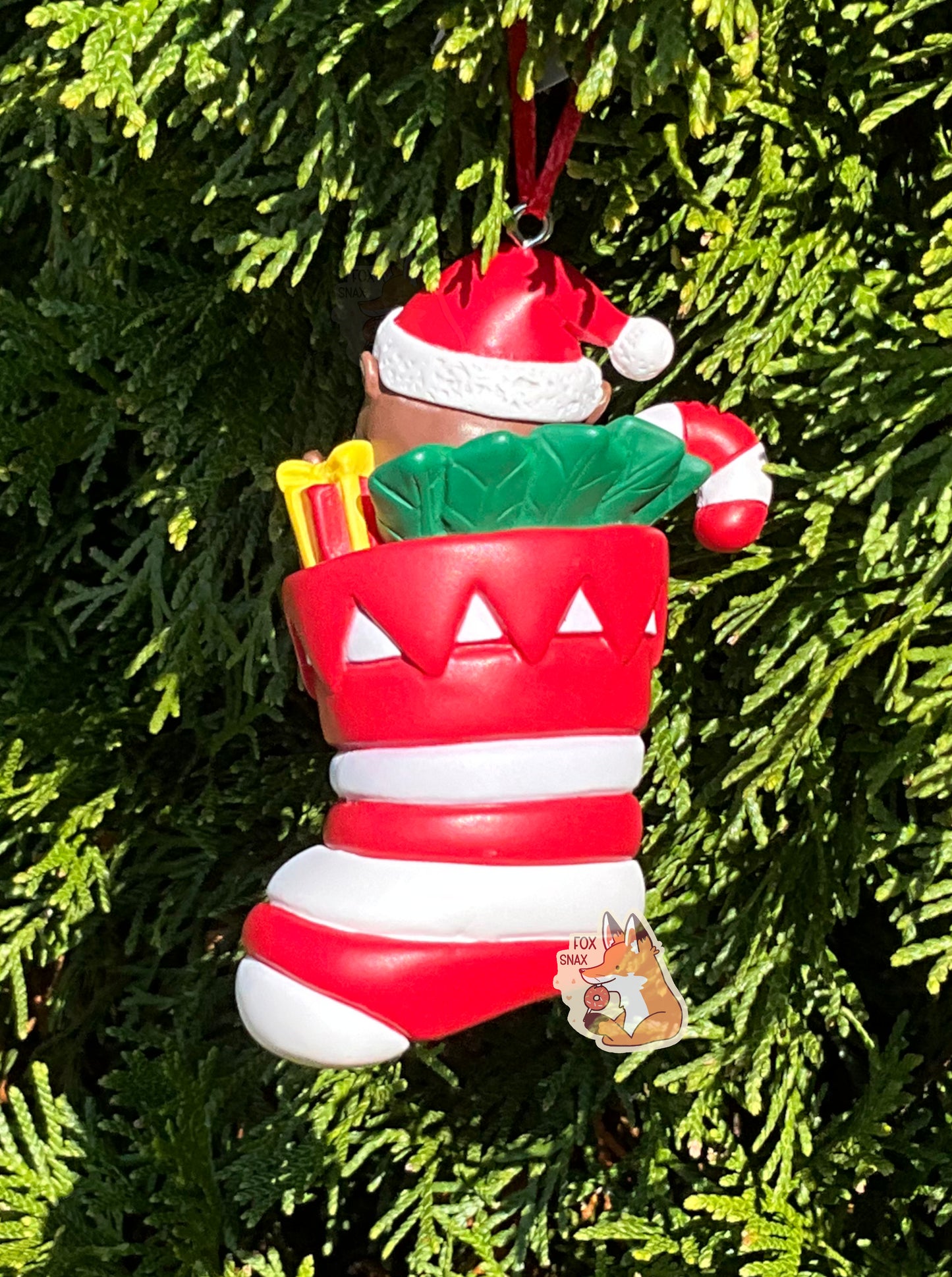 A picture of the back of a plastic ornament.  It has Buc-ee the Beaver, wearing a Santa hat and coat, sitting inside a red and white striped Christmas stocking.  In the stocking with Buc-ee are a red and white striped candy cane, bronze-colored bell, some sort of greenery, and a pack of Beaver Nuggets.  The stocking also has the Buc-ee's logo on it.  A small metal hook can be seen at the top of the ornament with an attached red ribbon.  The ornament is tied to a Christmas tree.