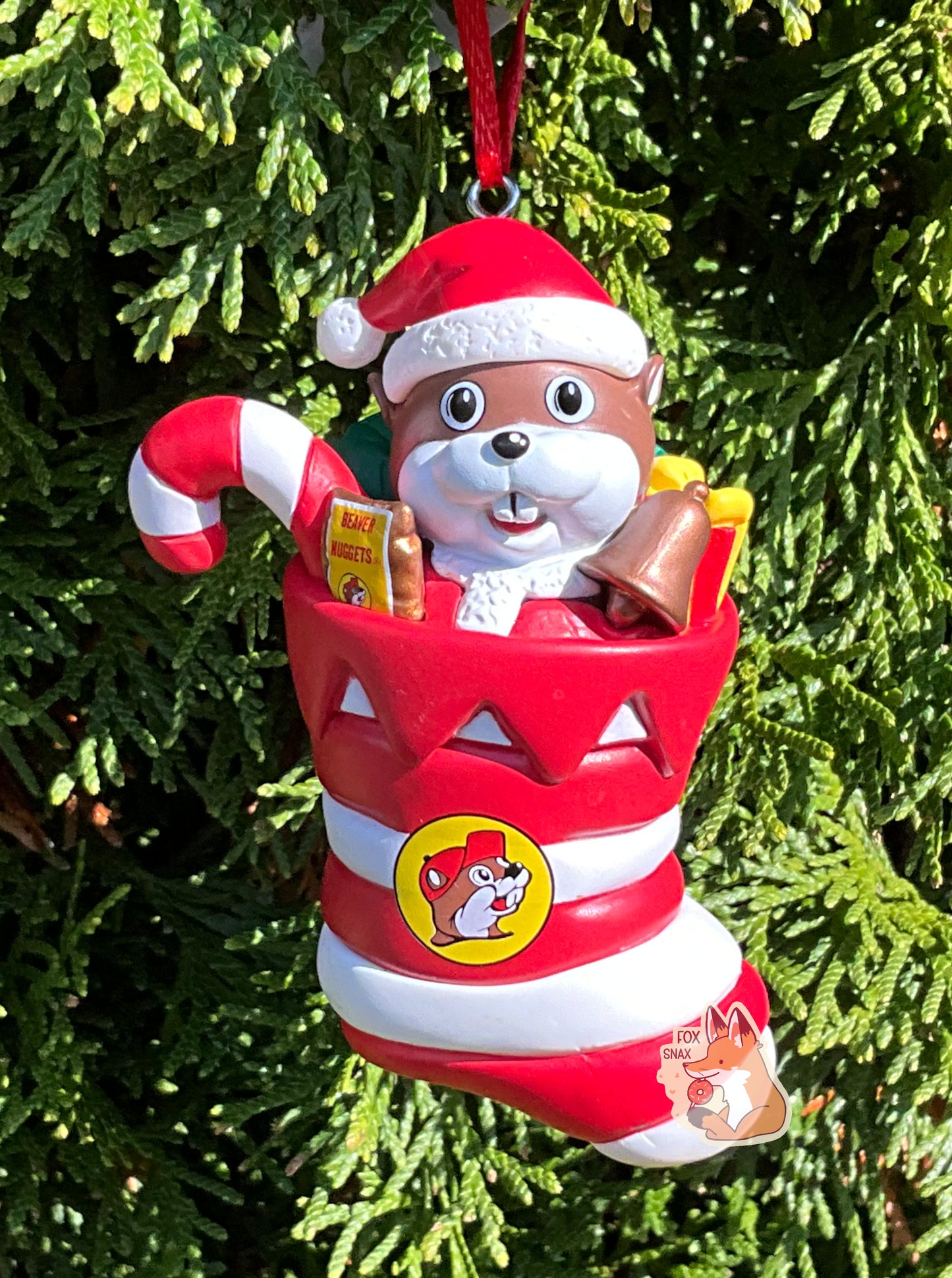 A picture of the front of a plastic ornament.  It has Buc-ee the Beaver, wearing a Santa hat and coat, sitting inside a red and white striped Christmas stocking.  In the stocking with Buc-ee are a red and white striped candy cane, bronze-colored bell, and a pack of Beaver Nuggets.  The stocking also has the Buc-ee's logo on it.  A small metal hook can be seen at the top of the ornament with an attached red ribbon.  The ornament is tied to a Christmas tree.