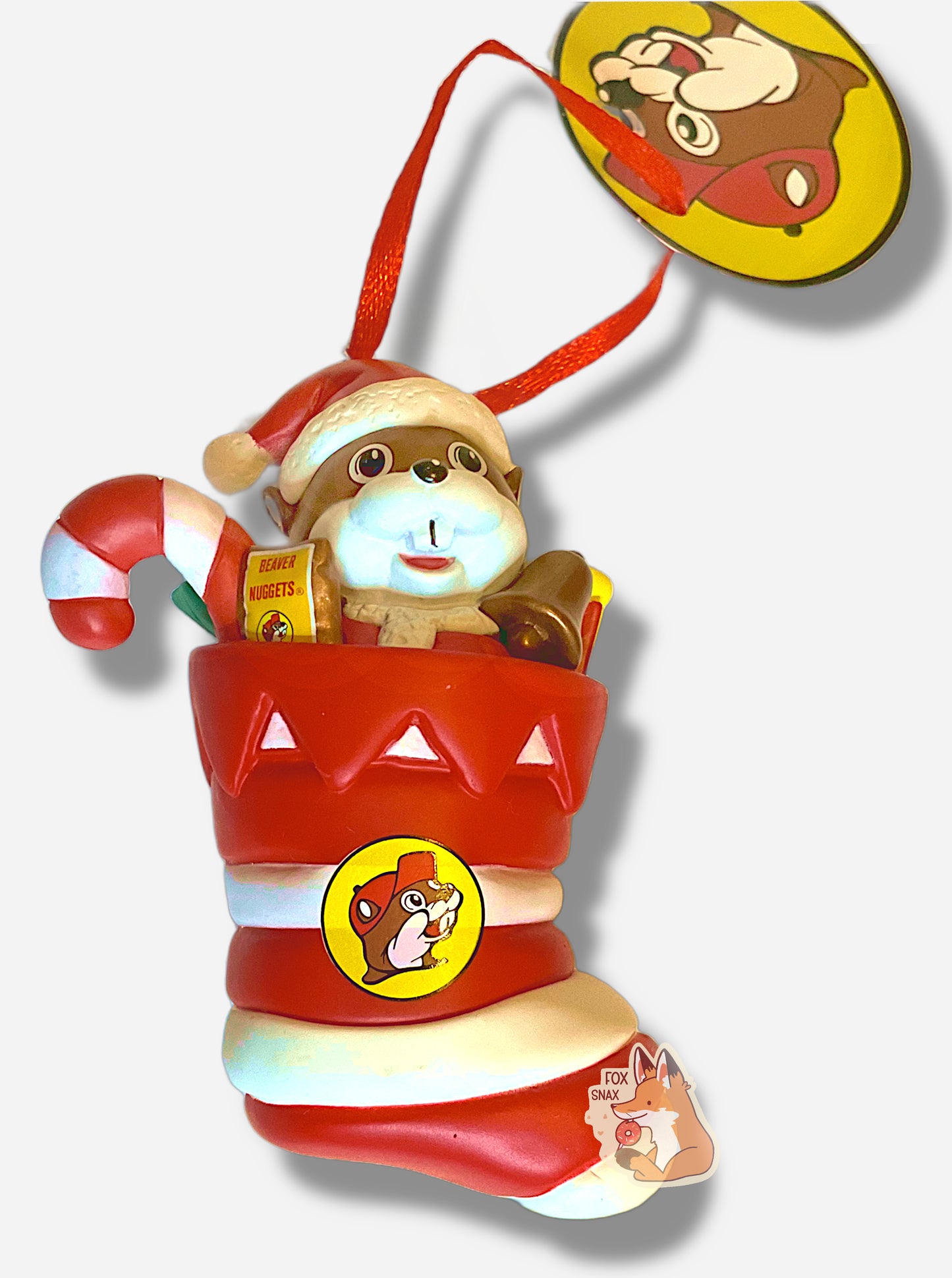 A picture of the front of a plastic ornament.  It has Buc-ee the Beaver, wearing a Santa hat and coat, sitting inside a red and white striped Christmas stocking.  In the stocking with Buc-ee are a red and white striped candy cane, bronze-colored bell, and a pack of Beaver Nuggets.  The stocking also has the Buc-ee's logo on it.  A small metal hook can be seen at the top of the ornament with an attached red ribbon. 