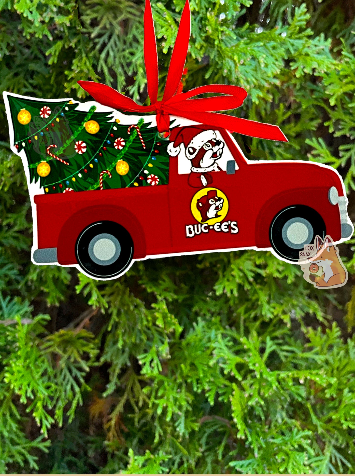 A picture of the front of a ceramic, mostly truck-shaped ornament, with a red ribbon at the top to hang it on a tree.  The ornament has a red old-style pickup truck, with Buc-ee The Beaver driving, with his paw hanging out the window.  Buc-ee is wearing a Santa hat.  The pickup truck's bed has a well decorated Christmas tree sitting in the bed, partially tipped over.  Candy canes, ornaments, colorful lights, and green garland decorate the tree. This ornament is hanging from an evergreen tree.