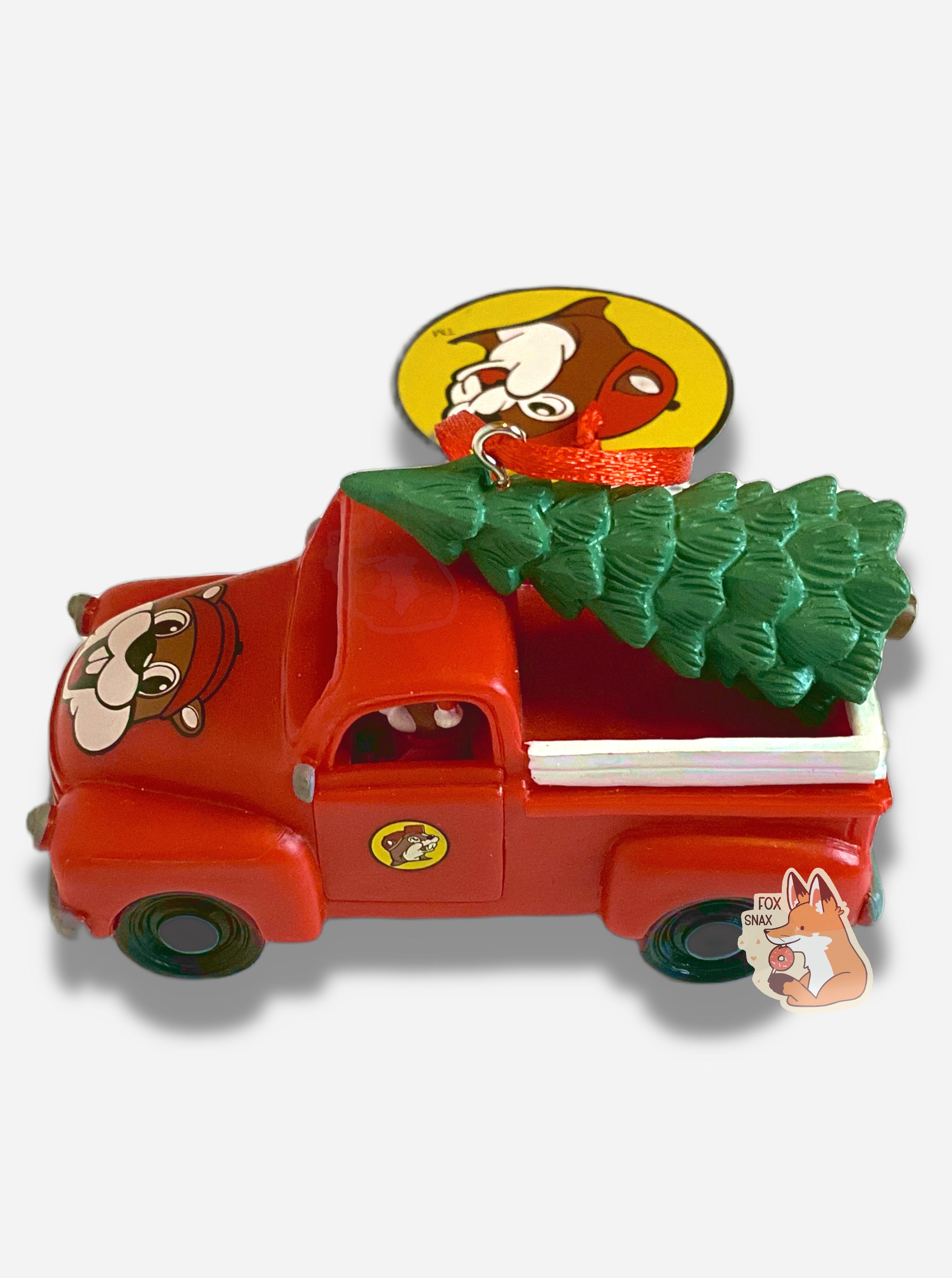 A picture of the side of a bright, plastic, Christmas ornament.  The ornament is of Buc-ee's the Beaver -  a smiling beaver wearing a red Christmas stocking cap. Buc-ee is driving a red pickup truck, with a green pine tree in the bed. 