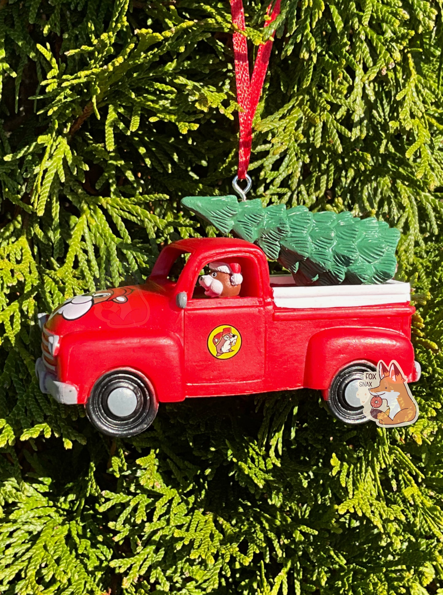 A picture of the side of a bright, plastic, Christmas ornament.  The ornament is of Buc-ee's the Beaver -  a smiling beaver wearing a red Christmas stocking cap. Buc-ee is driving a red pickup truck, with a green pine tree in the bed. A red ribbon pokes out from the top of the ornament's body, tying it to the green evergreen branches of a pretty tree in the background.