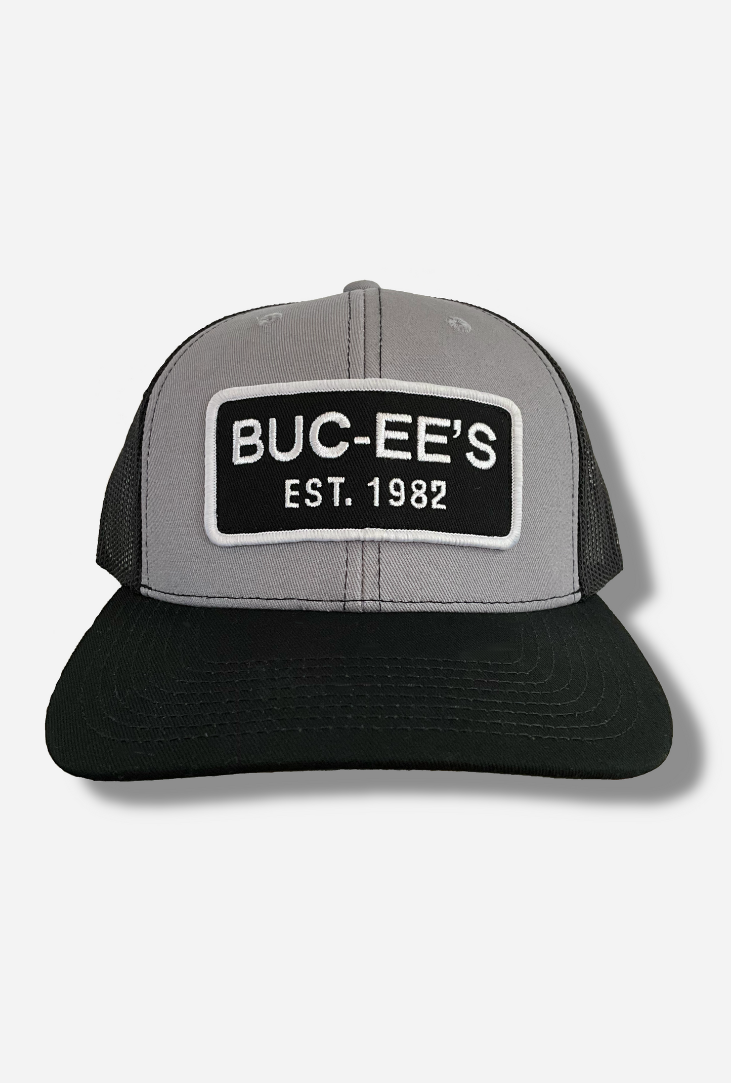 A picture of a grey and black ball cap.   The front of the cap is grey, and has a large white and black rectangular patch that reads BUC-EE'S, and underneath that, EST. 1982.  The brim of the cap is black.  The back and sides of the cap are a black plastic mesh.