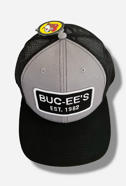 A picture of a grey and black ball cap.   The front of the cap is grey, and has a large white and black rectangular patch that reads BUC-EE'S, and underneath that, EST. 1982.  The brim of the cap is black.  The back and sides of the cap are a black plastic mesh.  A paper label is attached to the top of the cap; the label is shaped like the Buc-ee's logo.