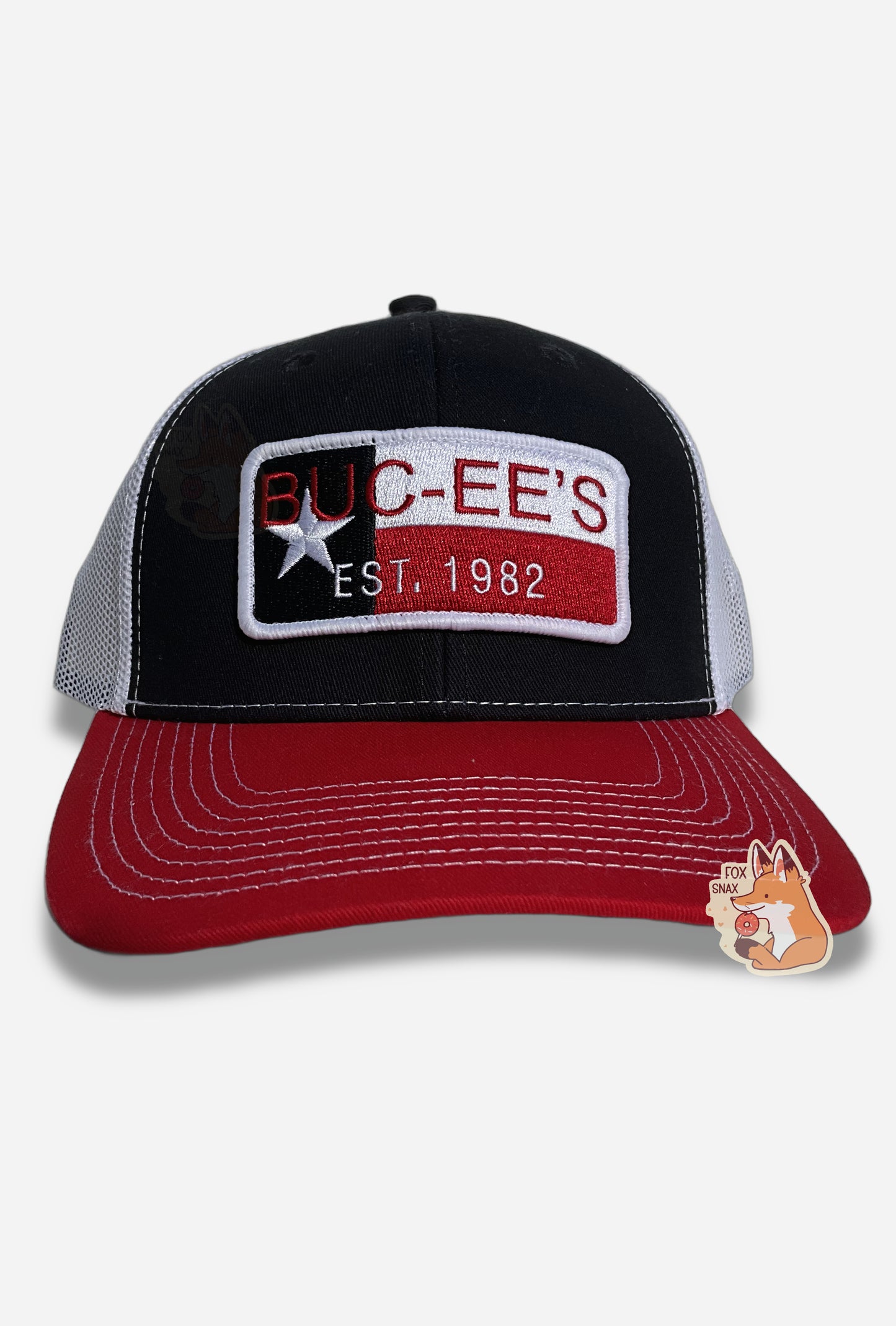 A picture of a red, white, and blue ball cap.   The front of the cap is dark blue, and has a large patch that reads BUC-EE'S in red, and underneath that in smaller white text, EST. 1982. The patch has a single white star, and blue, white, and red ines.  The brim of the cap is red.  The back and sides of the cap are a white plastic mesh.  