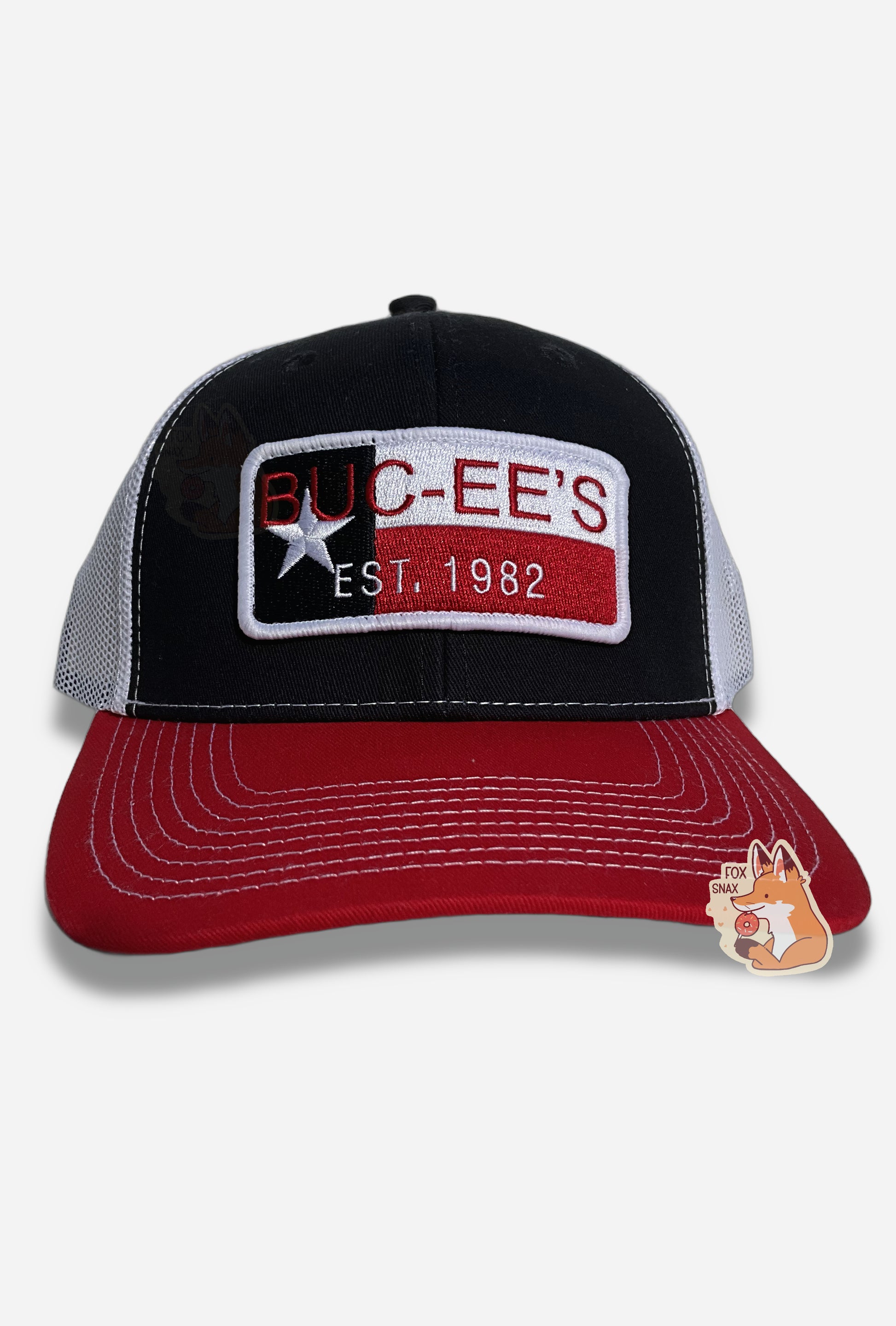 A picture of a red, white, and blue ball cap.   The front of the cap is dark blue, and has a large patch that reads BUC-EE'S in red, and underneath that in smaller white text, EST. 1982. The patch has a single white star, and blue, white, and red ines.  The brim of the cap is red.  The back and sides of the cap are a white plastic mesh.  