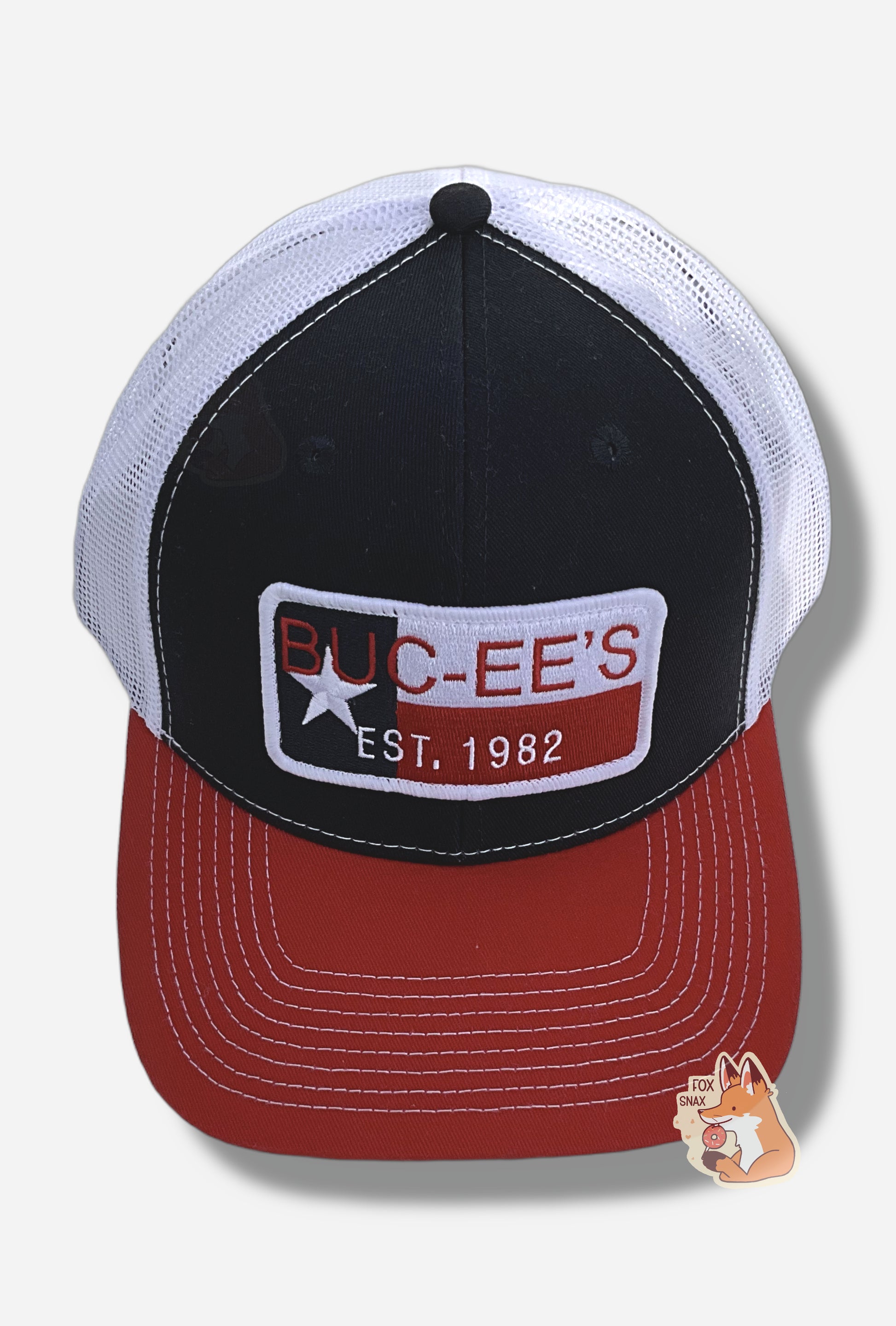 A three-quarters view of a red, white, and blue ball cap.   The front of the cap is dark blue, and has a large patch that reads BUC-EE'S in red, and underneath that in smaller white text, EST. 1982. The patch has a single white star, and blue, white, and red ines.  The brim of the cap is red.  The back and sides of the cap are a white plastic mesh.  