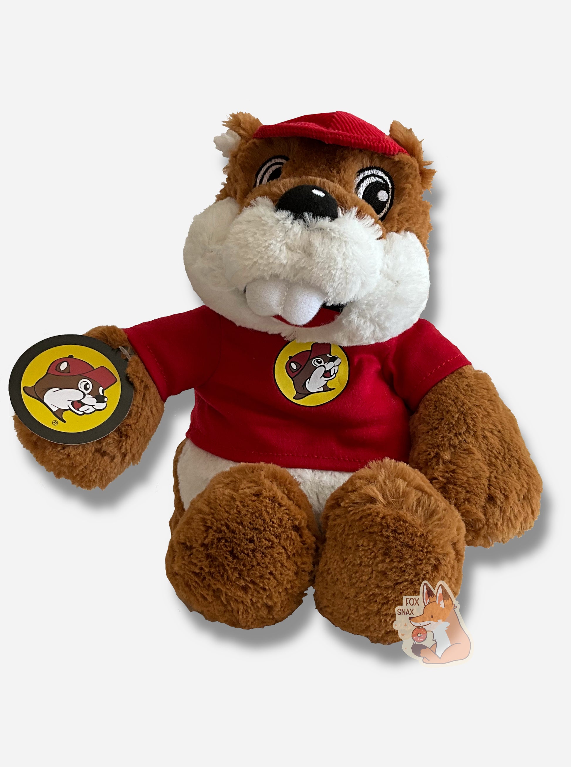 A picture of an adorable Buc-ee the Beaver warmie sitting down.  Buc-ee has brown and white fur, and is wearing a red cap.  His shirt is a red shirt, with a Buc-ee's logo on it.