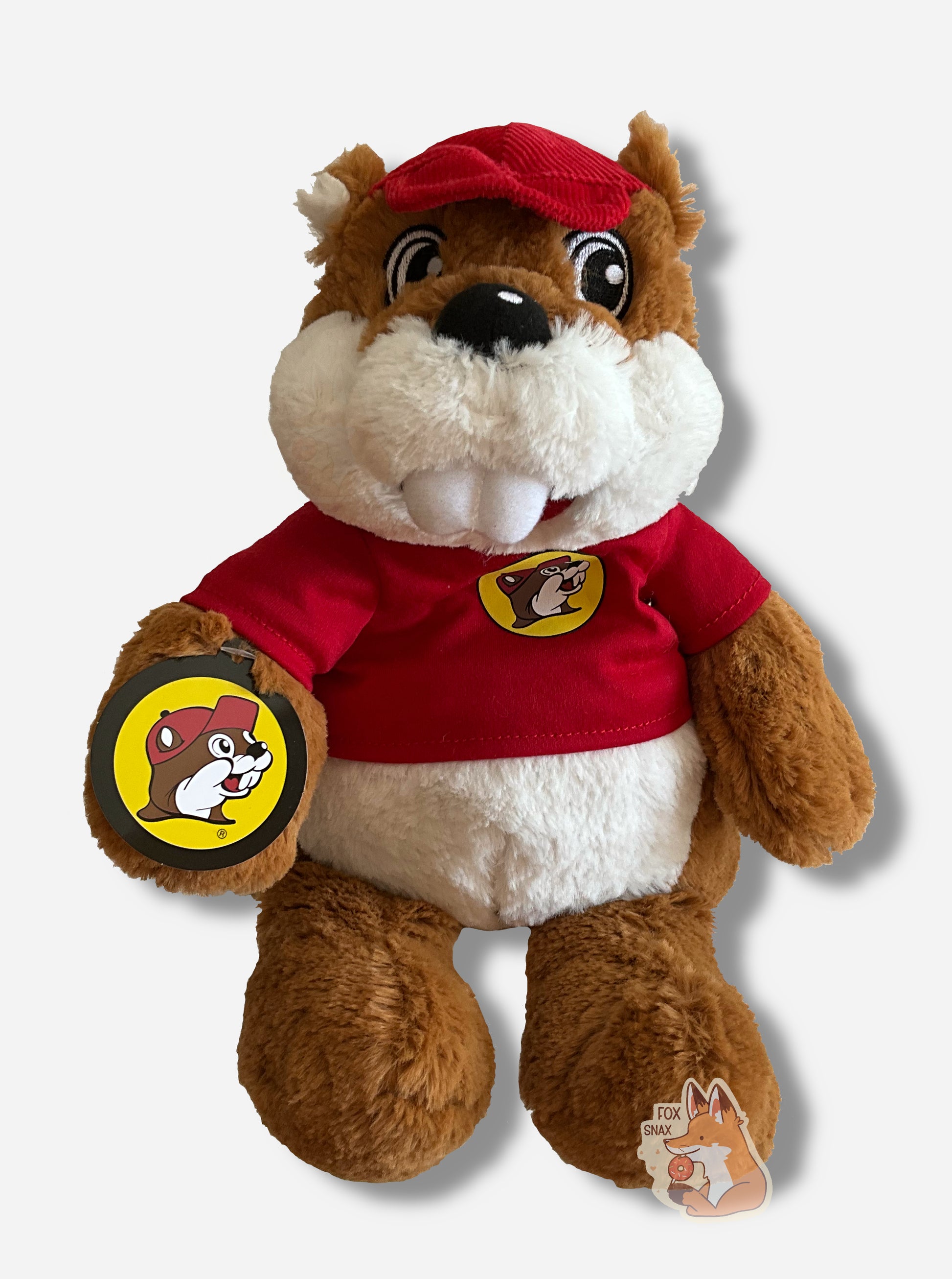 A picture of an adorable Buc-ee the Beaver warmie sitting.  Buc-ee has brown and white fur, and is wearing a red cap.  His shirt is a red shirt, with a Buc-ee's logo on it.