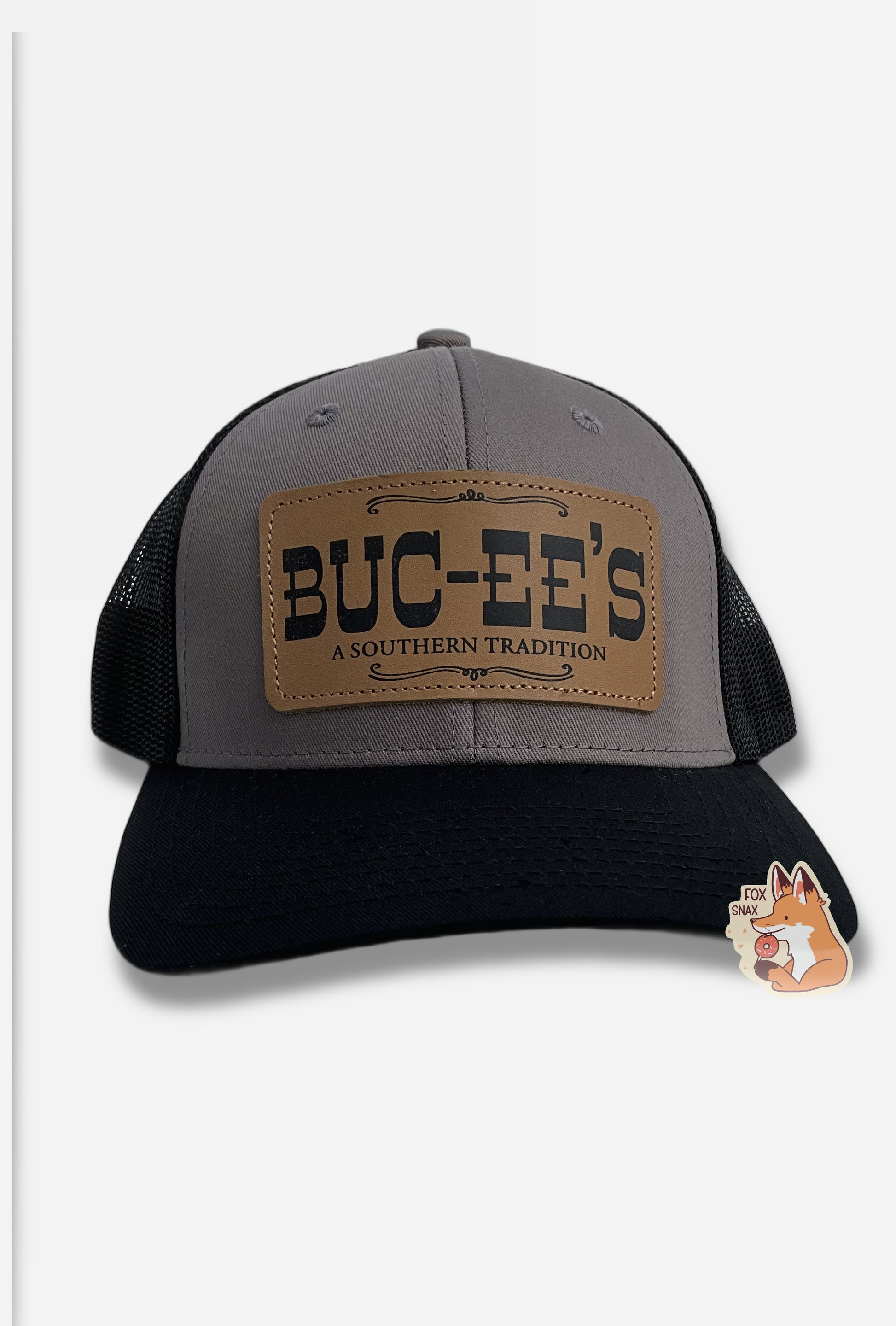 A picture of a grey and black ball cap.   The front of the cap is grey, and has a large rectangular leather patch that reads BUC-EE'S, and underneath that, A SOUTHERN TRADITION.  The brim of the cap is black.  The back and sides of the cap are a black plastic mesh.