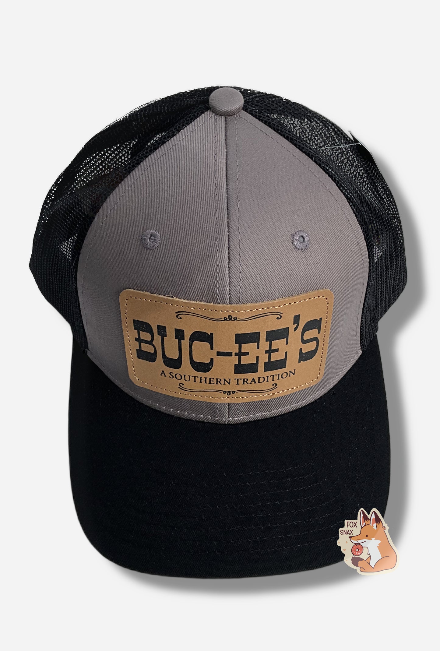 A picture of a grey and black ball cap.   The front of the cap is grey, and has a large rectangular leather patch that reads BUC-EE'S, and underneath that in much smaller text, A SOUTHERN TRADITION.  The brim of the cap is black.  The back and sides of the cap are a black plastic mesh.  A paper label is attached to the top of the cap; it reads TOP GRAIN LEATHER PATCH.